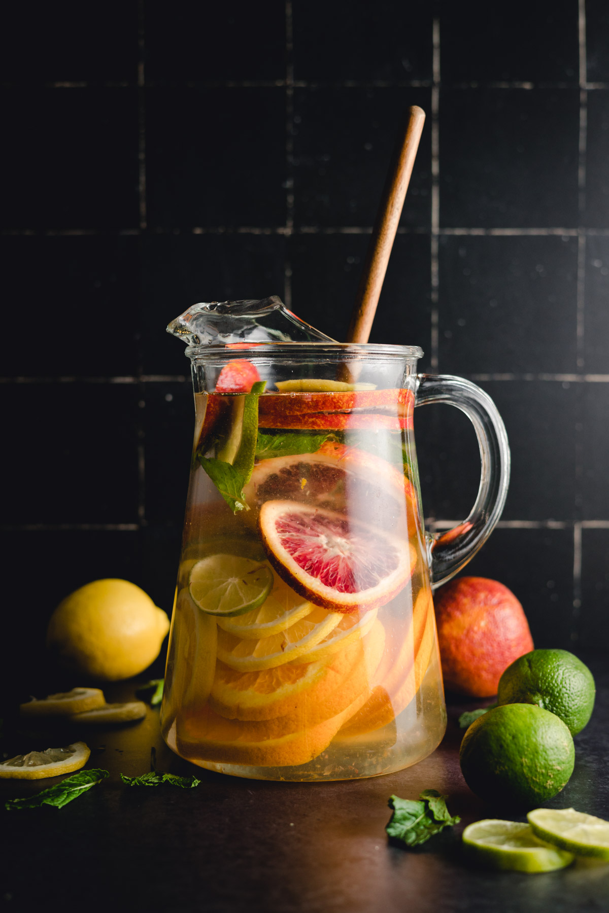 A clear glass pitcher filled with water, sliced citrus fruits including blood oranges, limes, and a lemon, with a wooden stirring stick. Whole fruits are scattered around the base.