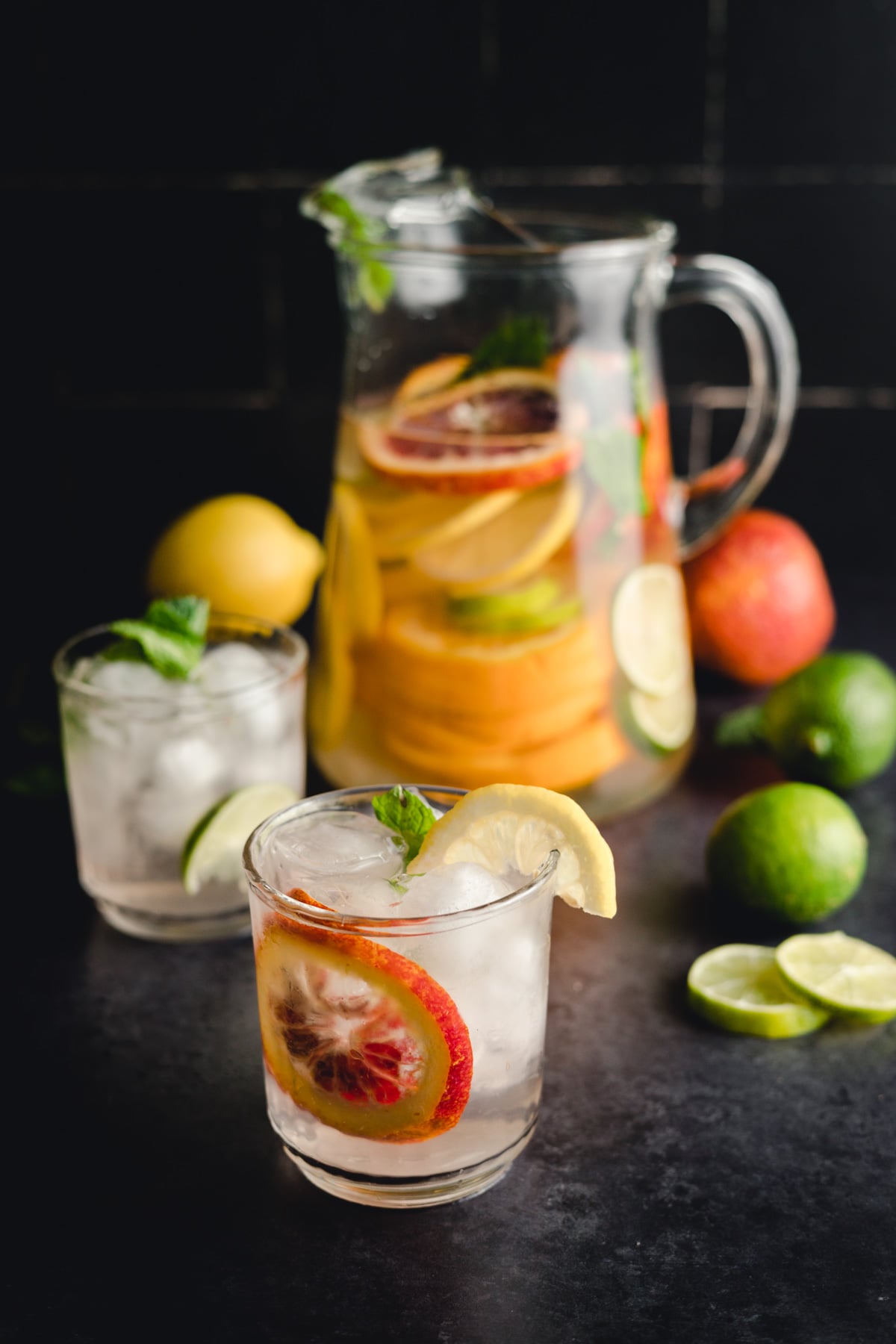 Glass pitcher and two glasses filled with a citrus-infused drink with ice cubes, garnished with lemon, lime, and mint; whole fruits and sliced citrus in the background on a dark surface.
