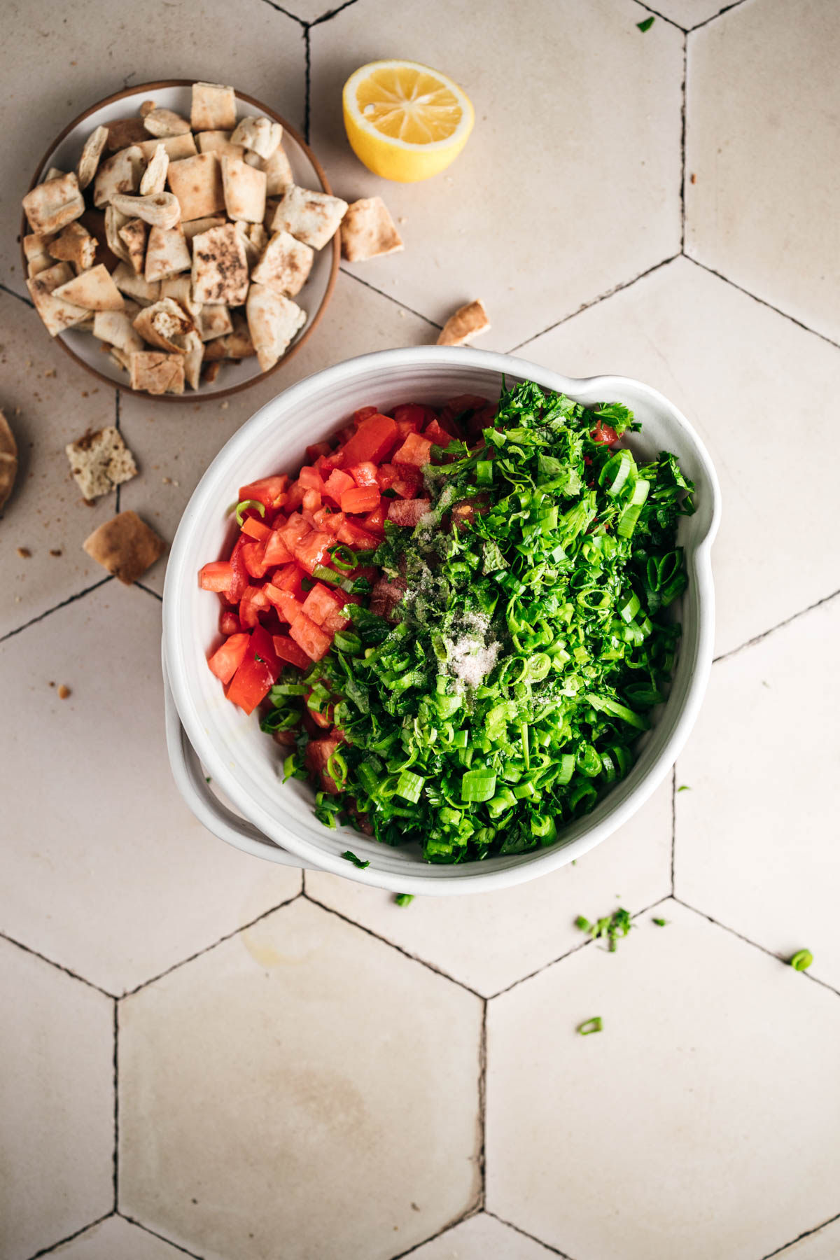 A bowl with chopped tomatoes, parsley, and other vegetables on a tiled surface, accompanied by a small dish of chopped pita bread and a half lemon.
