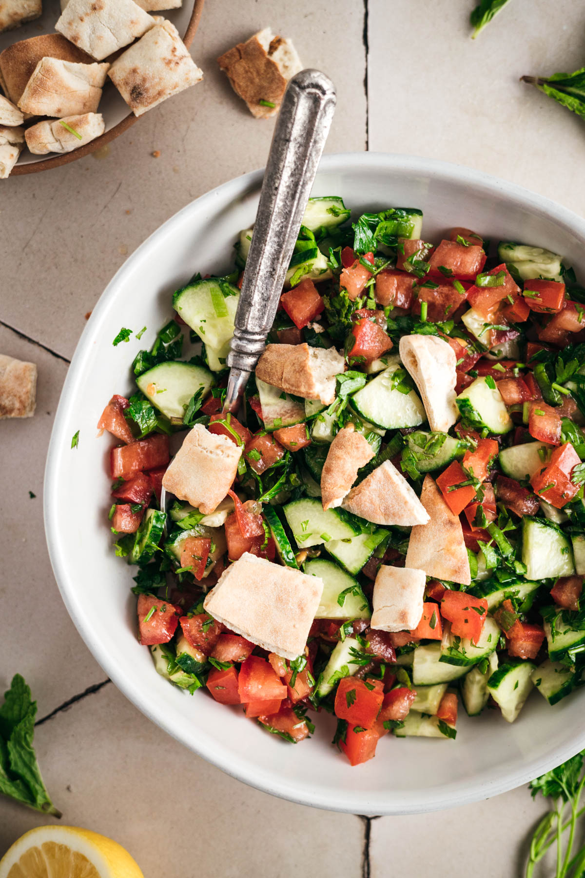 A white bowl filled with a salad of chopped tomatoes, cucumbers, parsley, and pita bread pieces. A metal spoon is placed in the bowl. Pita bread pieces and fresh herbs are scattered around the bowl.