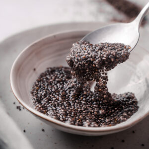 A close-up of a spoon lifting soaked chia seeds, resembling a chia egg, from a bowl on a white plate. The seeds have a gelatinous texture.