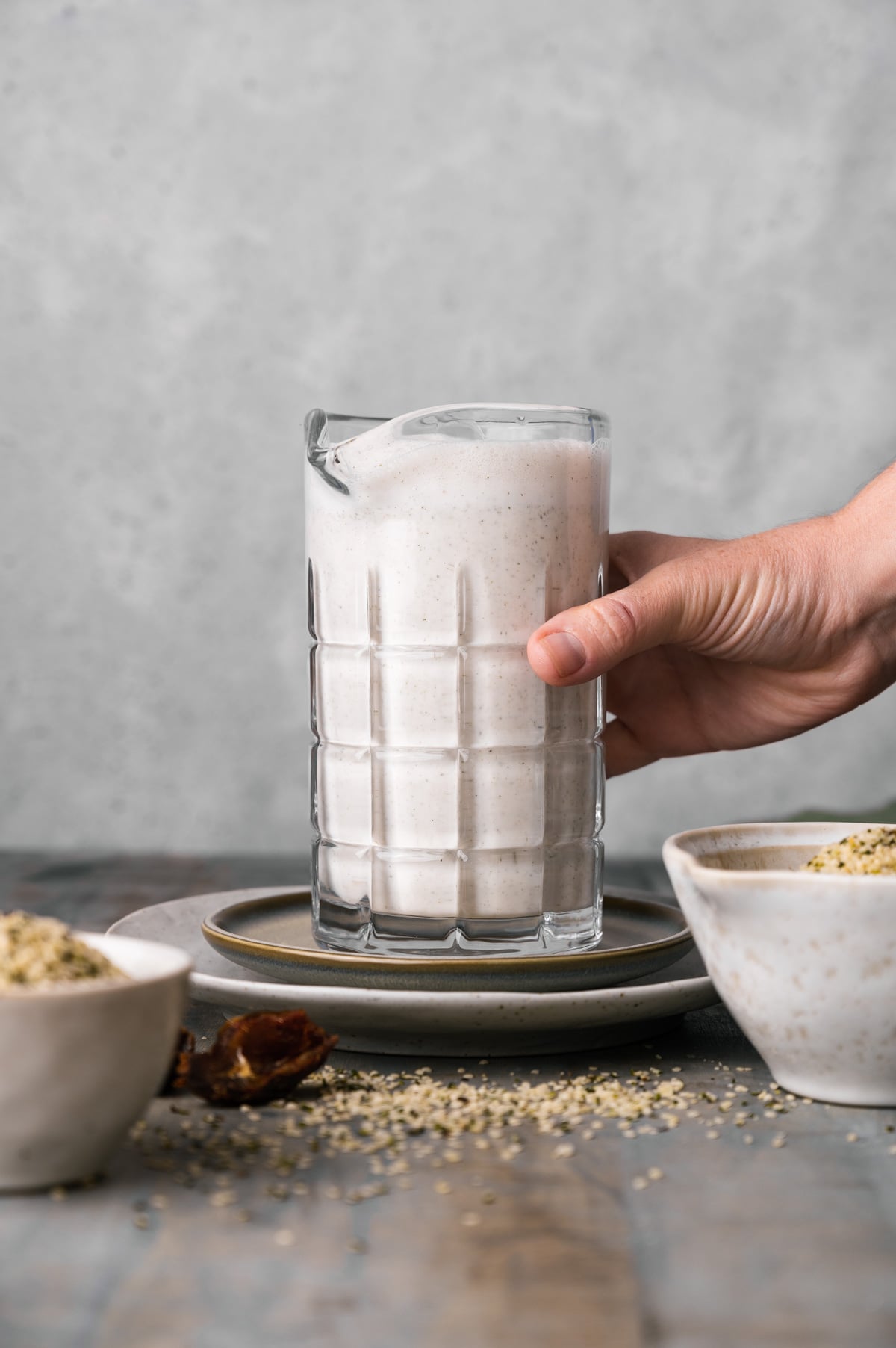 A hand holding a glass pitcher filled with creamy hemp milk, placed on a stack of two plates. Surrounding the pitcher are bowls with seeds and a smooth, grey surface in the background.