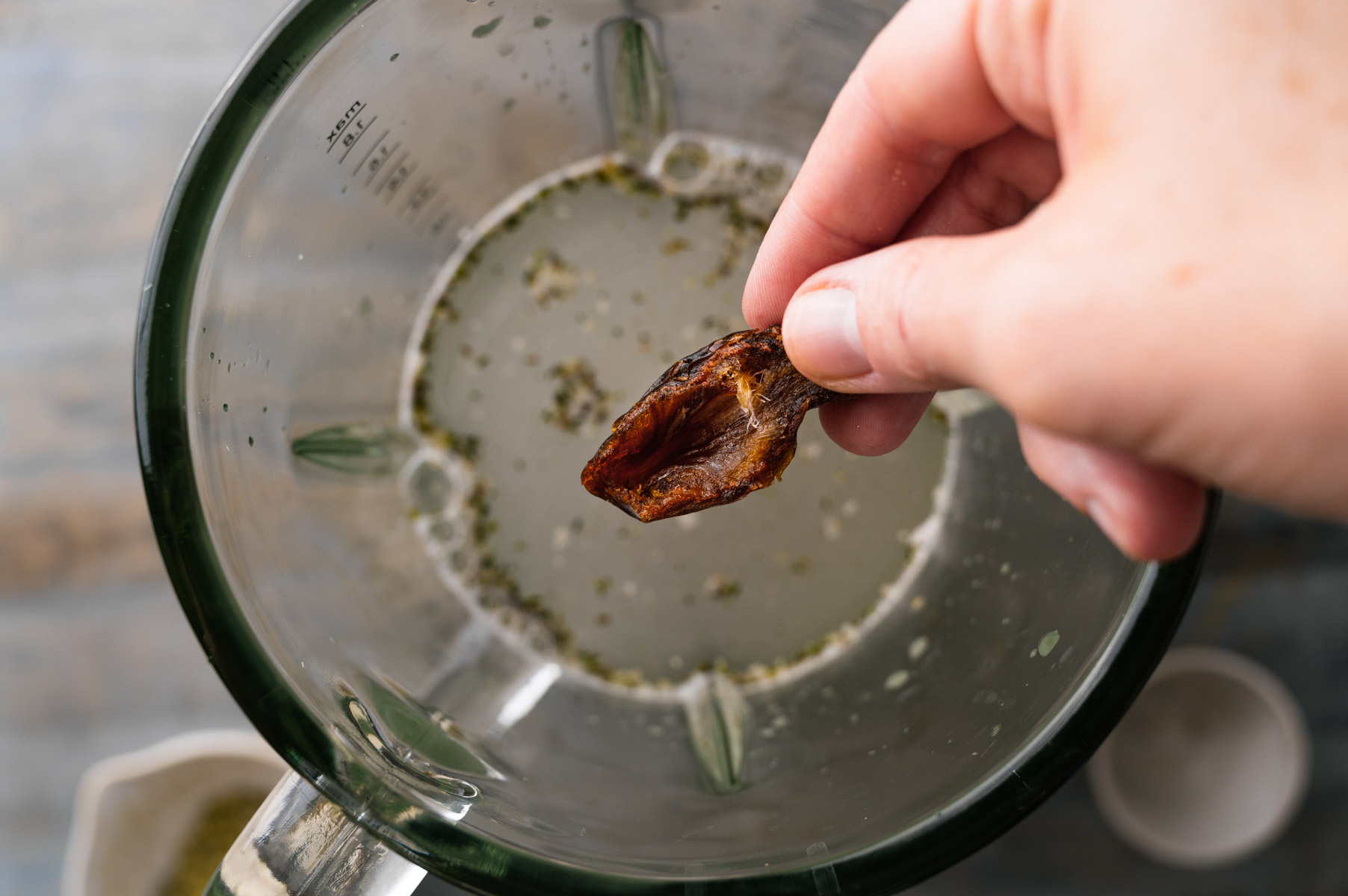A hand holds a date over an open blender containing liquid and other ingredients.