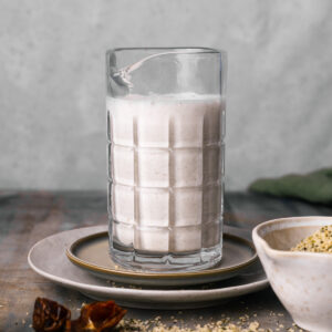 A frothy glass of plant-based milk sits on a plate; a bowl containing seeds and some spilled seeds are visible on a wooden surface nearby.