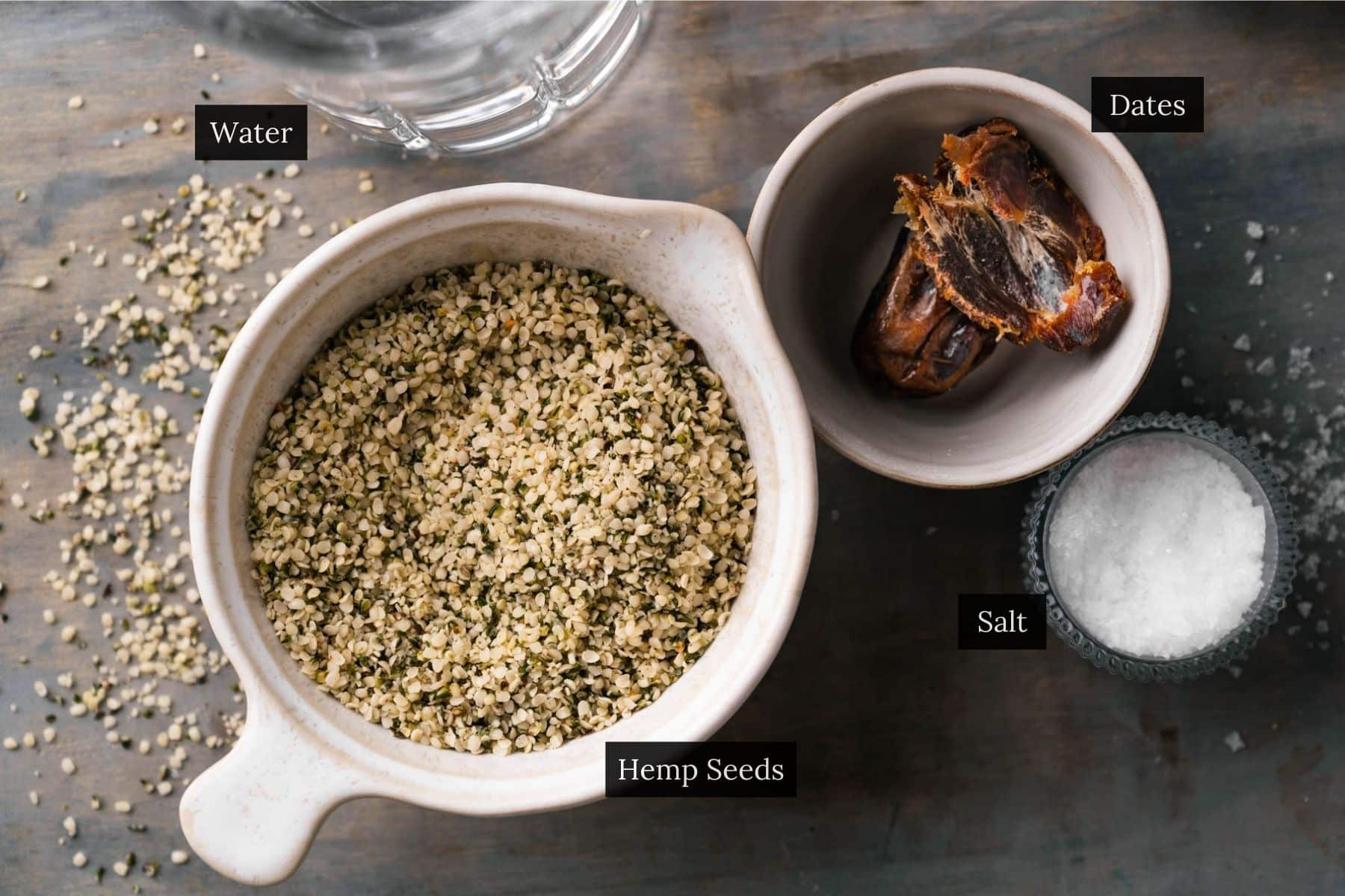 A top view of bowls containing hemp seeds, dates, and salt, alongside a pouring glass ready to create creamy hemp milk.