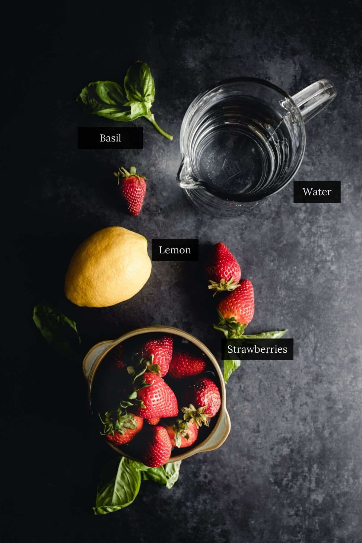 A glass jug of water, basil leaves, a lemon, and a bowl of strawberries arranged on a dark surface table.