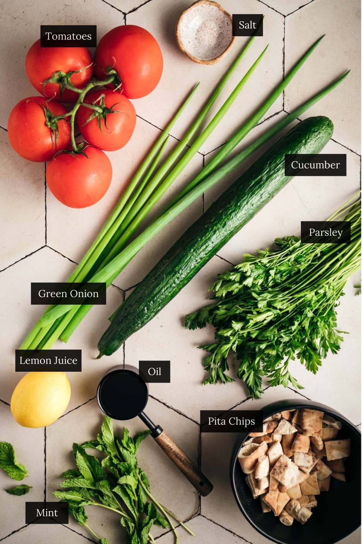 A Mediterranean medley on a tiled surface: ripe tomatoes, crisp cucumber, fresh green onions, parsley, and mint mingle with zesty lemon, salt, oil, and crunchy pita chips.