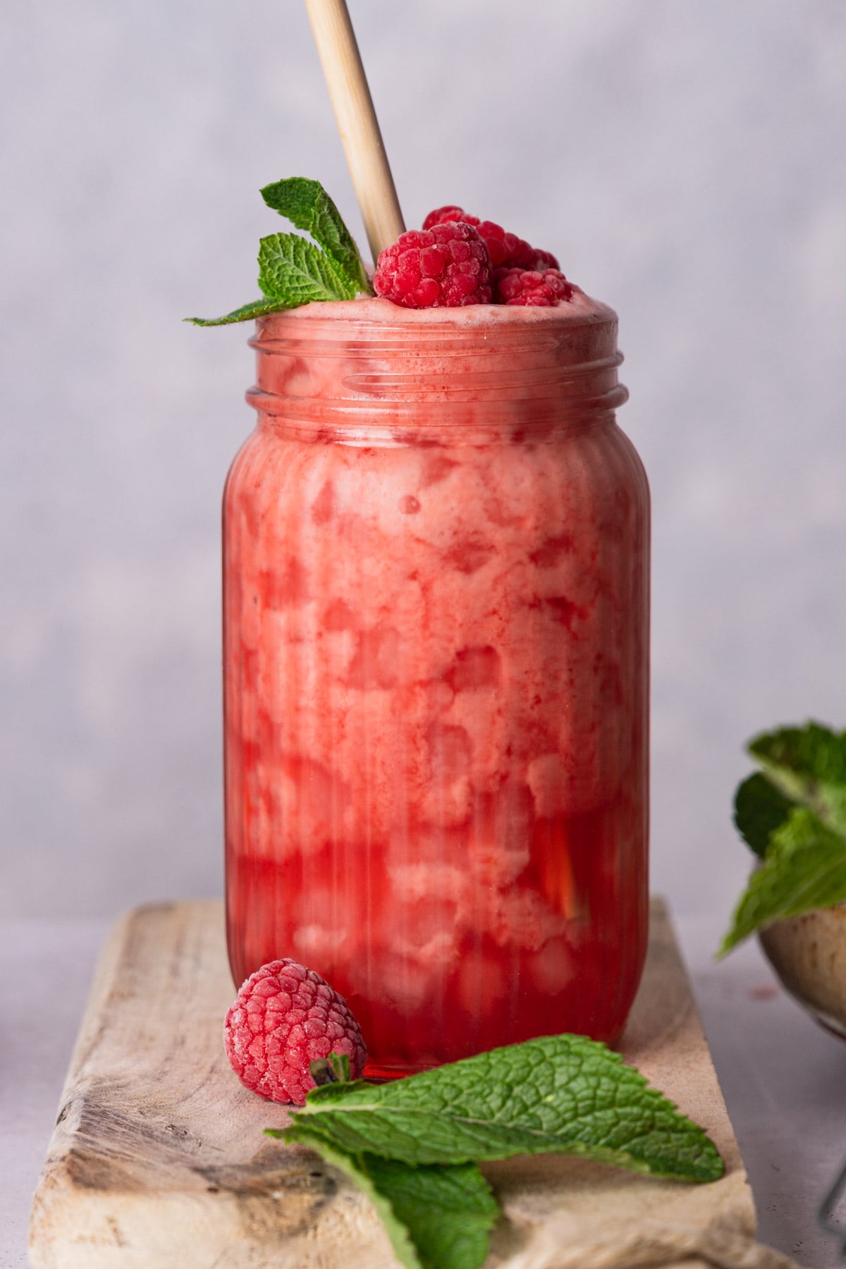 A frozen raspberry lemonade in a mason jar with a bamboo straw, topped with raspberries and a sprig of fresh mint, placed on a wooden surface with additional mint leaves and a raspberry beside it.