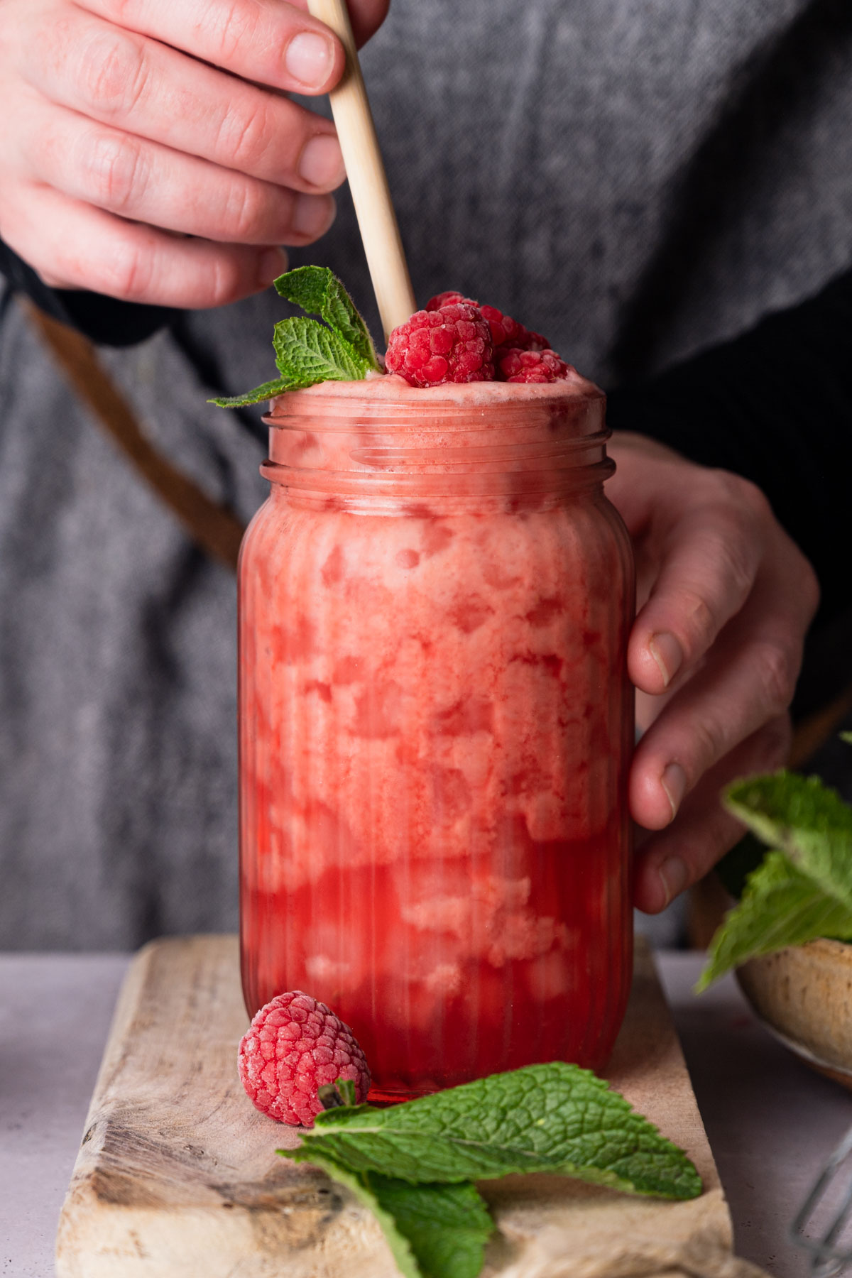 A person stirs a raspberry lemonade drink topped with raspberries and mint in a mason jar placed on a wooden board.