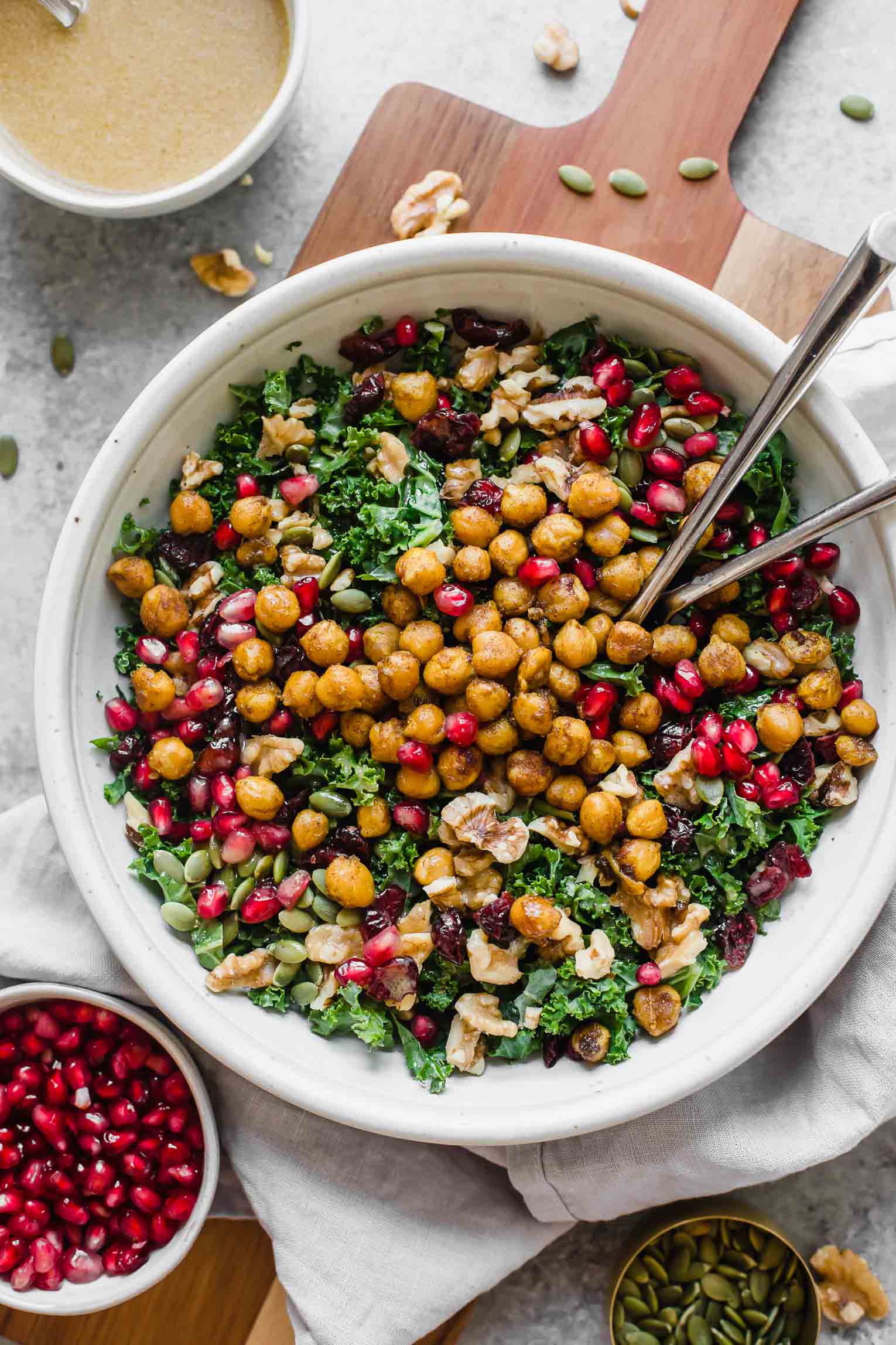 A bowl of kale salad topped with roasted chickpeas, pomegranate seeds, nuts, and seeds, accompanied by a small bowl of pomegranate seeds, another with seeds, and a cup of dressing on the side. For a complete meal idea from Tasty Recipes, consider pairing it with a warm butternut squash soup.