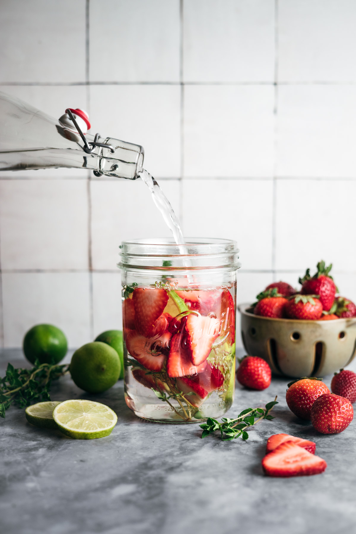 A glass bottle pours water into a jar filled with strawberries, lime slices, and herbs on a counter. A bowl of strawberries, lime, and herbs are scattered around.