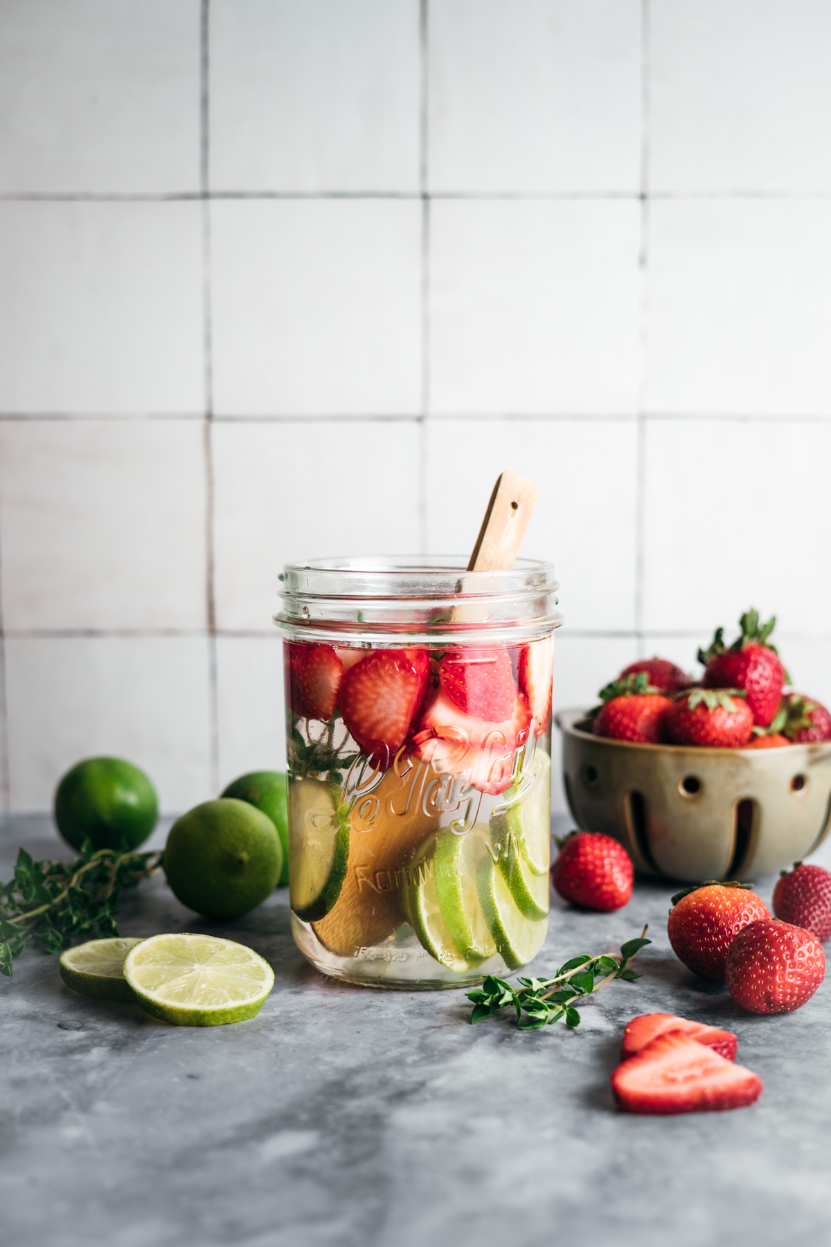 A mason jar filled with water, sliced limes, strawberries, and mint leaves, with a wooden spoon. Limes and a bowl of strawberries are placed nearby on a gray surface against a tiled backdrop.