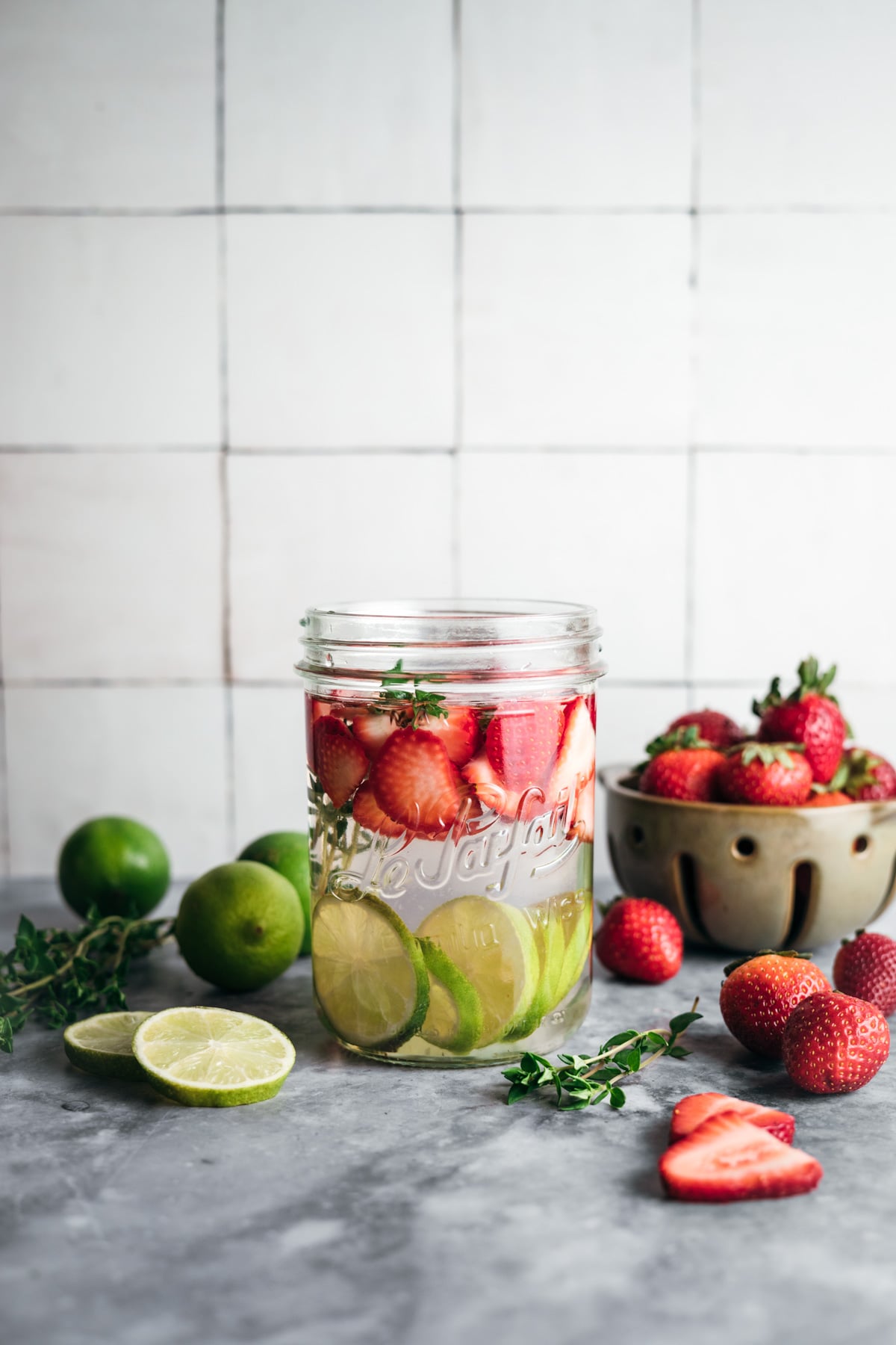 A mason jar filled with water, sliced strawberries, and limes sits on a countertop. Fresh strawberries, limes, and greens are arranged around the jar. A bowl of strawberries is in the background.
