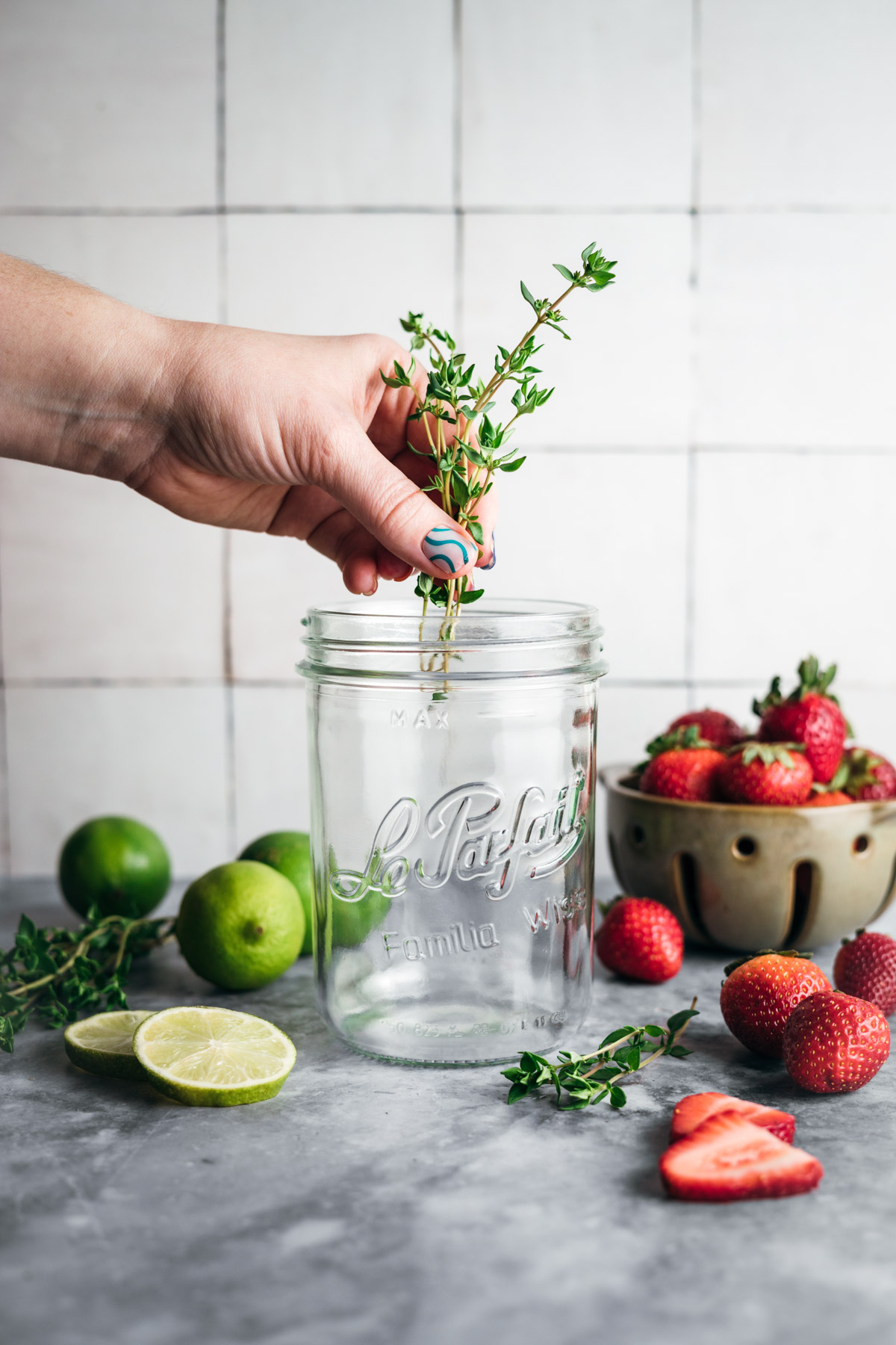 A hand places sprigs of thyme into an empty glass jar. Fresh strawberries, lime halves, whole limes, and more sprigs of thyme are scattered on a gray countertop. A ceramic bowl holds more strawberries.