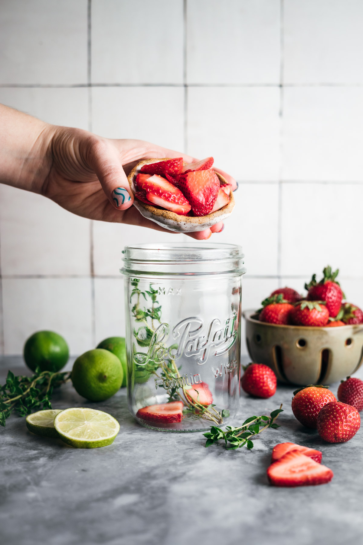 A hand is placing strawberries on top of a mason jar, with sliced limes, strawberries, and herbs on the gray surface below. A bowl of strawberries and some lime halves are in the background.