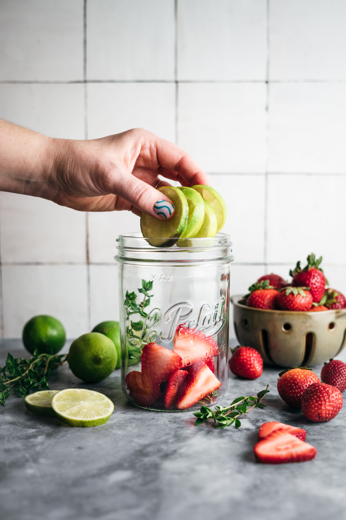 A hand places green apple slices into a mason jar filled with strawberries, surrounded by limes, cut fruit, and a bowl of strawberries on a countertop.