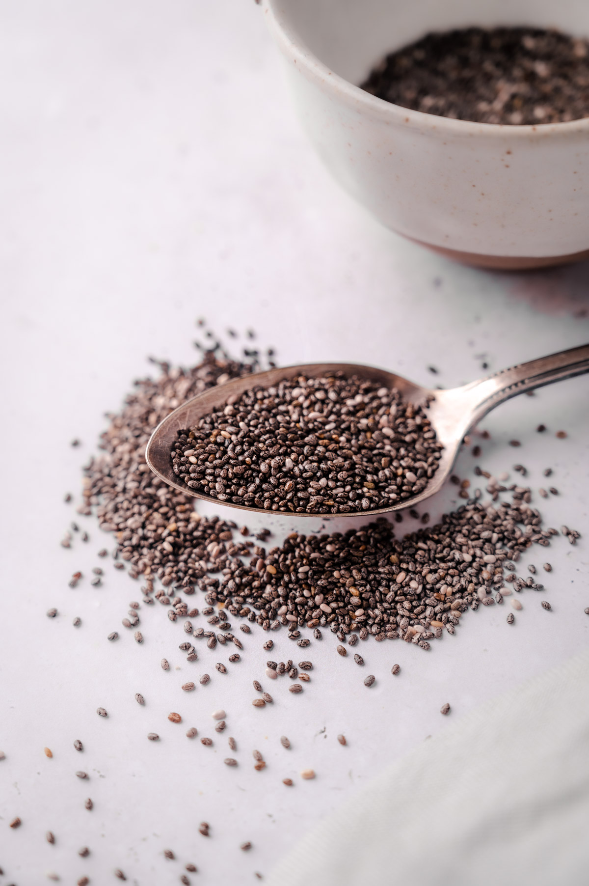 A spoon filled with chia seeds rests on a table with more chia seeds scattered and a bowl partially visible in the background.