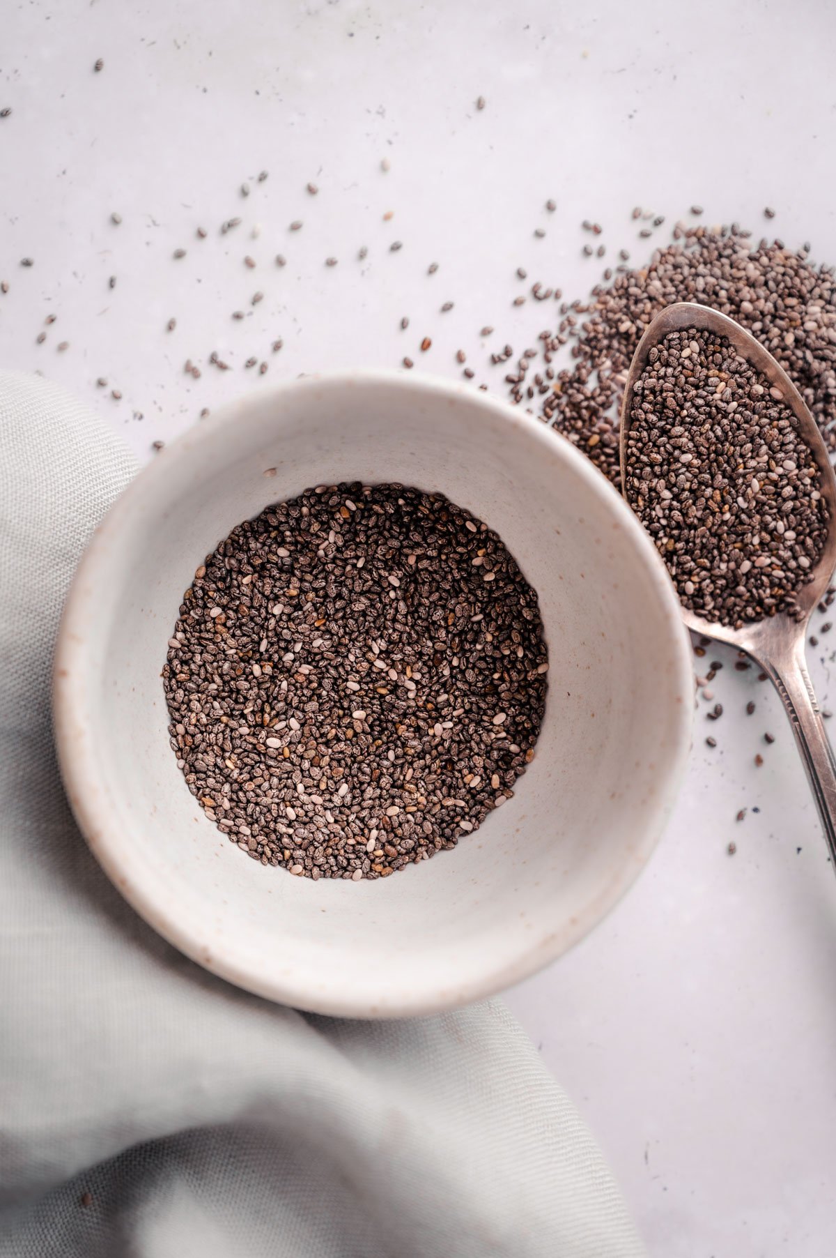 A white bowl filled with chia seeds next to a spoon with chia seeds on a light-colored surface. A cloth is partially visible to the left of the bowl.