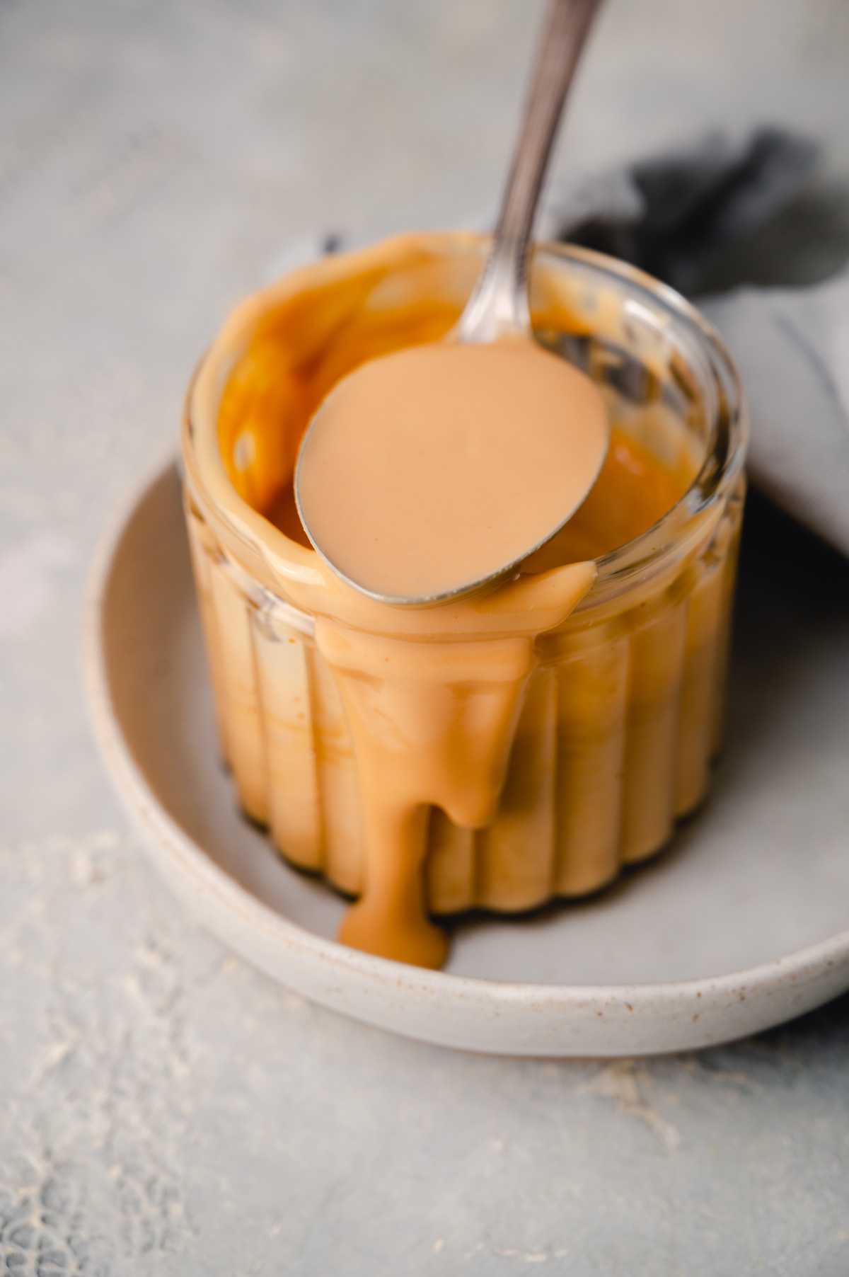 A spoon rests on top of a glass jar filled with almond butter, with it dripping onto a white plate underneath.
