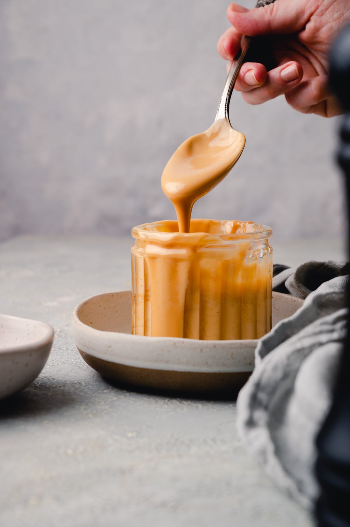 A hand holding a spoon with almond butter drizzling into a clear glass jar. 