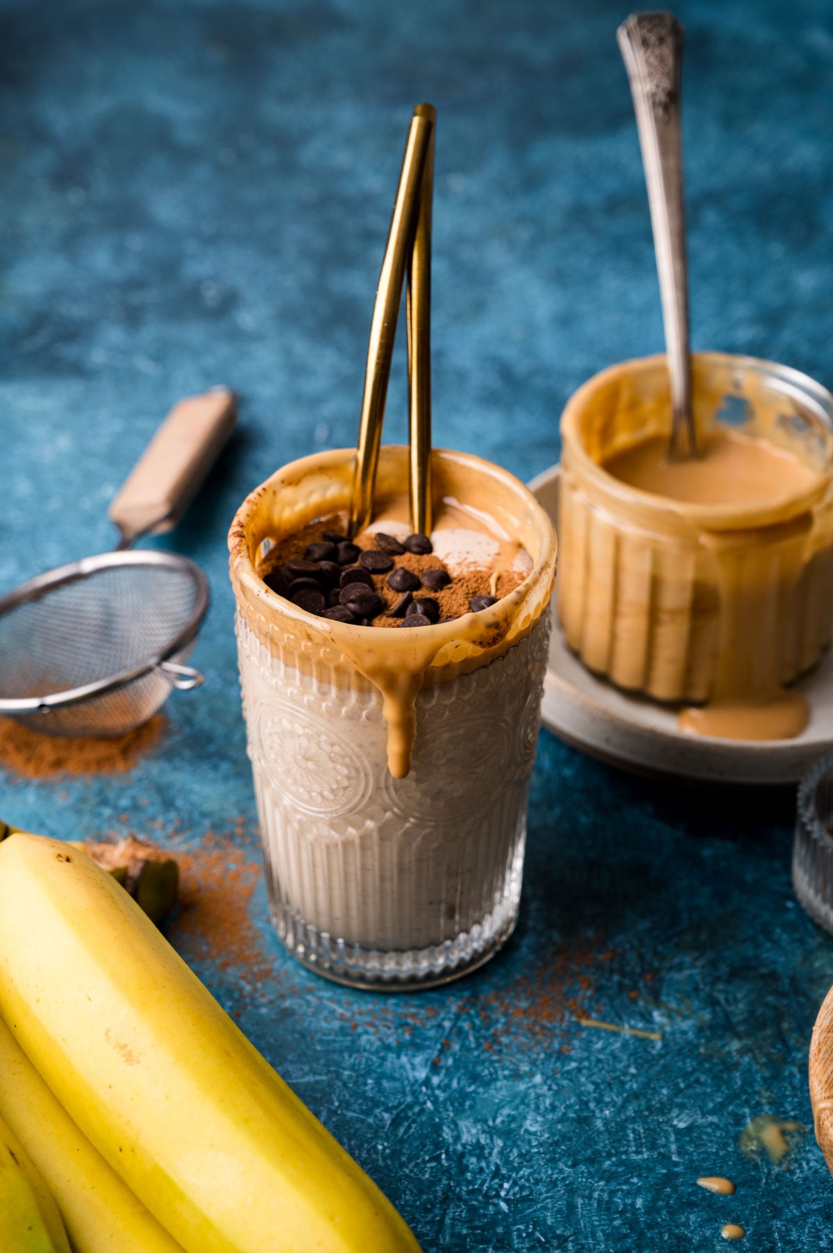A glass of smoothie topped with a thick layer of almond butter, chocolate chips, and crushed nuts. Bananas, a strainer, a jar of almond butter, and a spoon are on the table.
