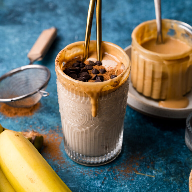 A glass of banana almond butter smoothie topped with chocolate chips and peanut butter, with bananas and a strainer in the background on a blue surface.
