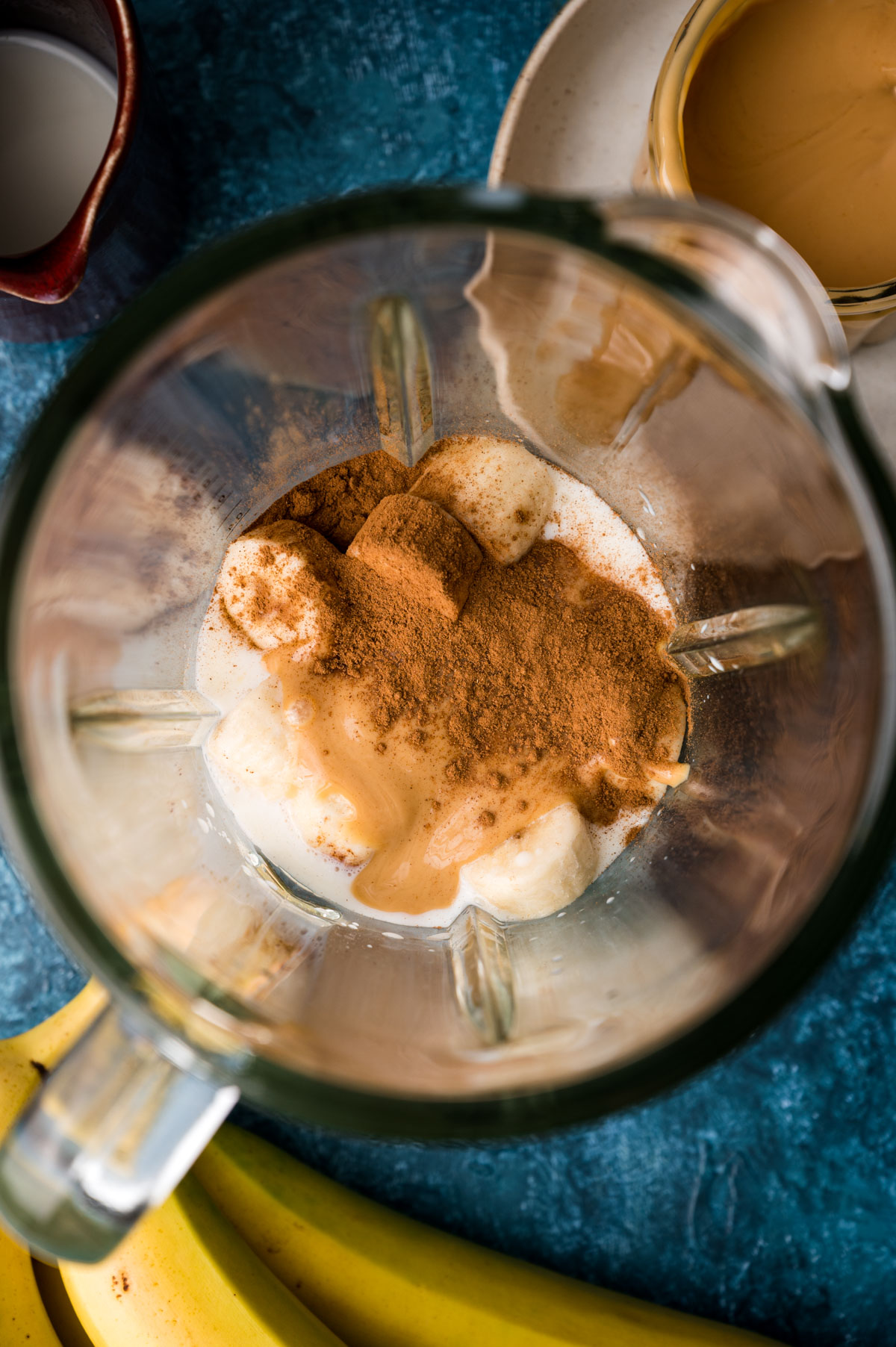 Blender with almond milk, almond butter, bananas, and cinnamon on a blue surface.