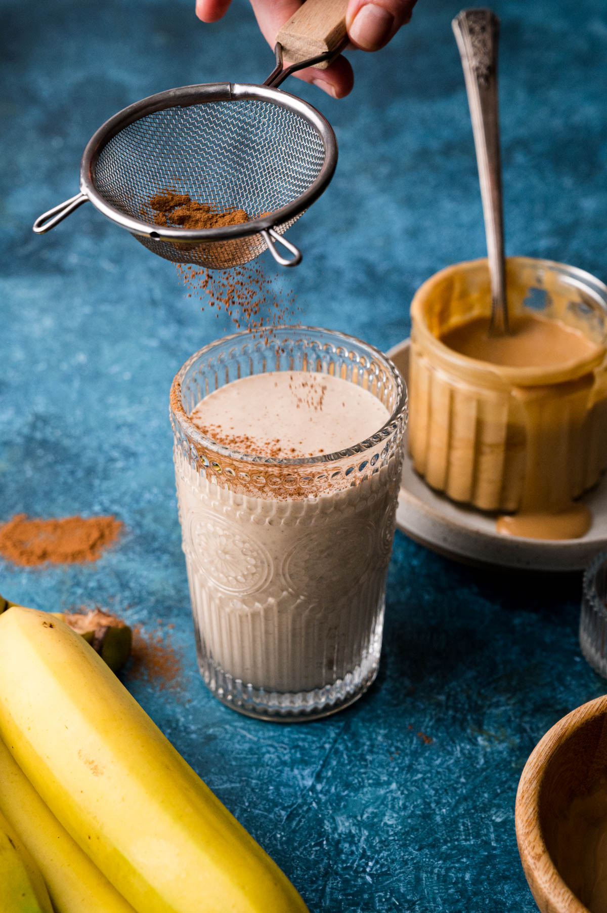 A glass of smoothie is being dusted with cinnamon through a sieve. Nearby are bananas and a jar of almond butter with a spoon in it on a blue surface.