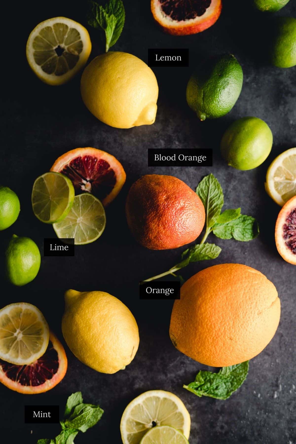 An assortment of citrus fruits labeled as lemon, blood orange, lime, and orange, with mint leaves scattered on a dark surface.