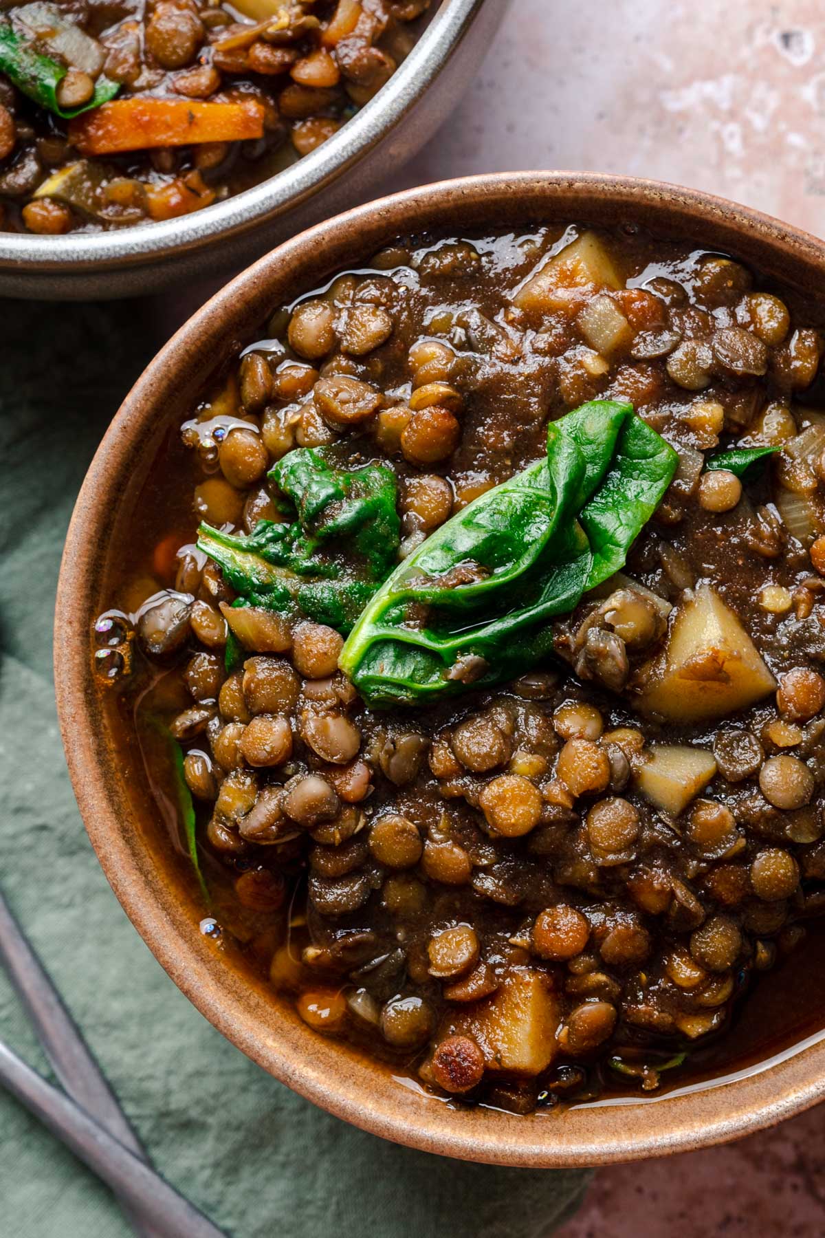 A bowl of lentil soup with spinach and chopped vegetables, placed on a tablecloth with a spoon nearby.