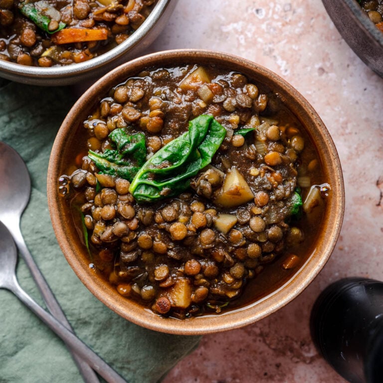 A bowl of lentil soup with spinach and diced potatoes on a textured surface, next to two spoons and a green napkin.