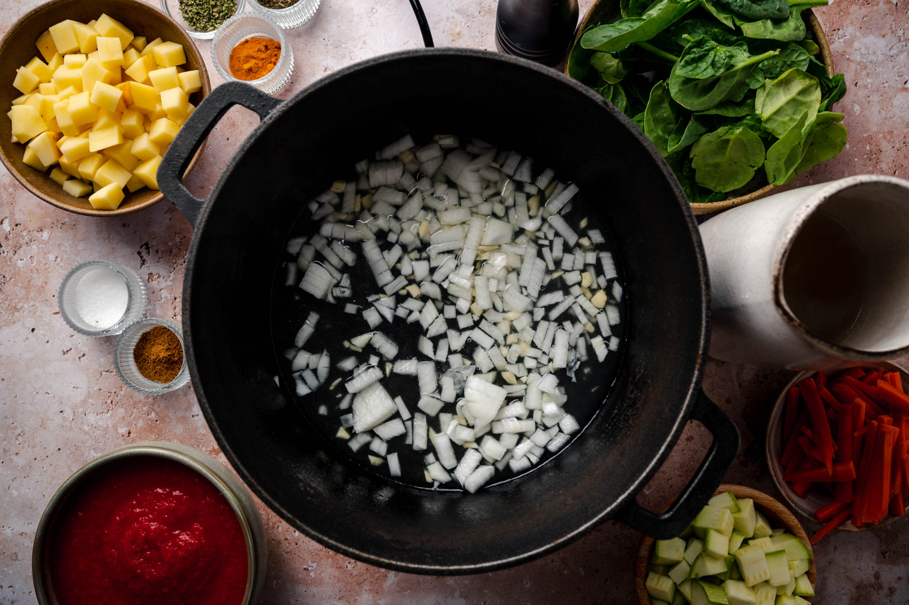 A black pot with diced onions at the center, surrounded by bowls containing diced potatoes, spinach leaves, carrot sticks, spices, salt, and tomato paste.