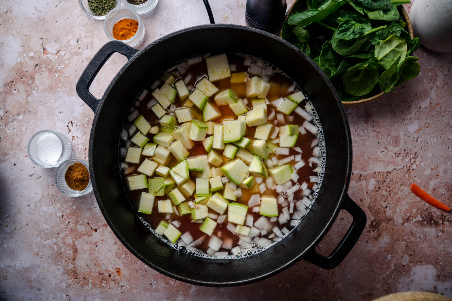 A Dutch oven containing diced vegetables and broth is placed on a marble countertop. Surrounding the pot are small bowls of spices, salt, and fresh spinach leaves.
