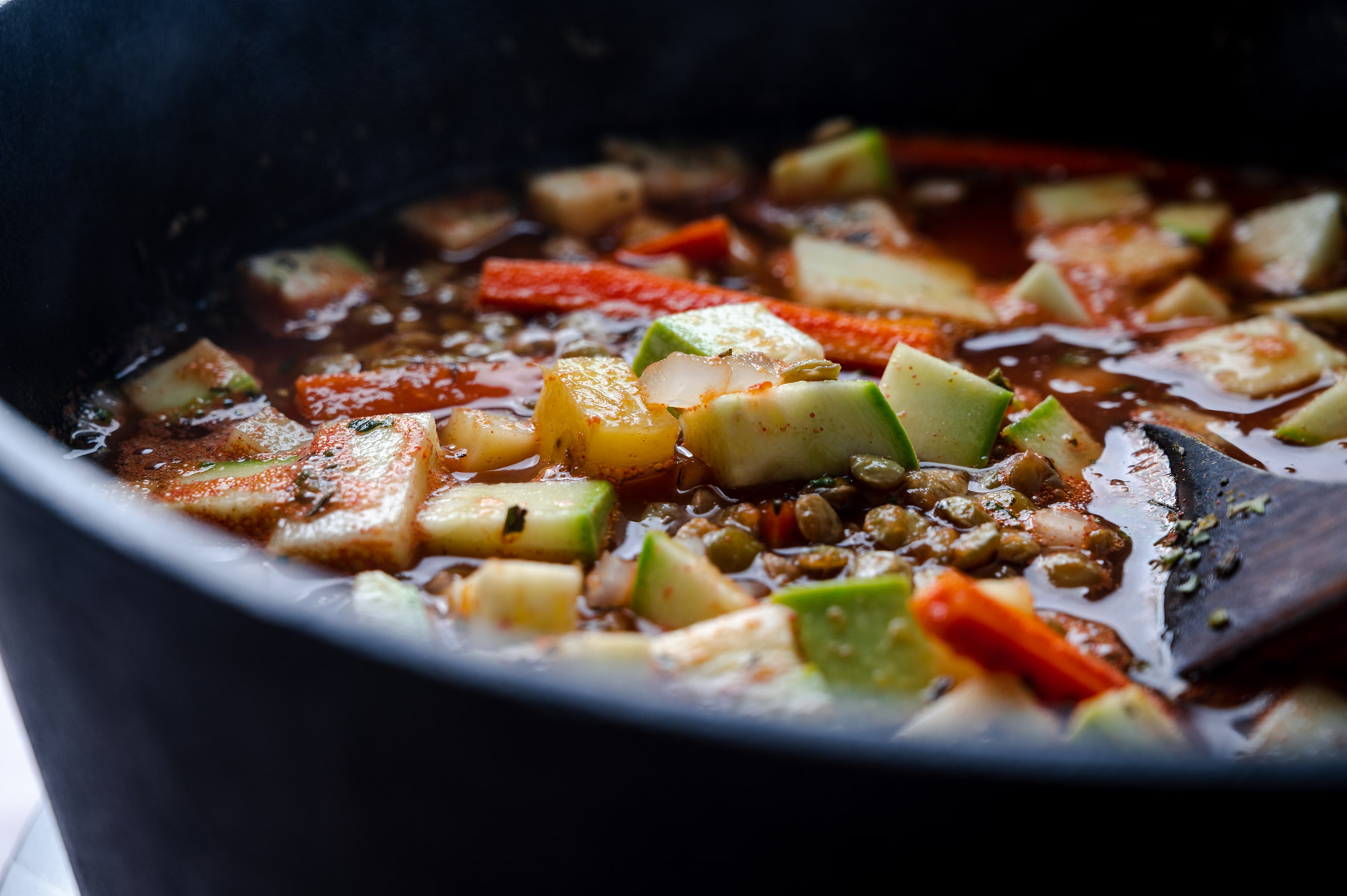 A close-up of a pot containing a simmering vegetable stew with chopped zucchini, carrots, bell peppers, lentils, and a wooden spoon partially visible in the broth.