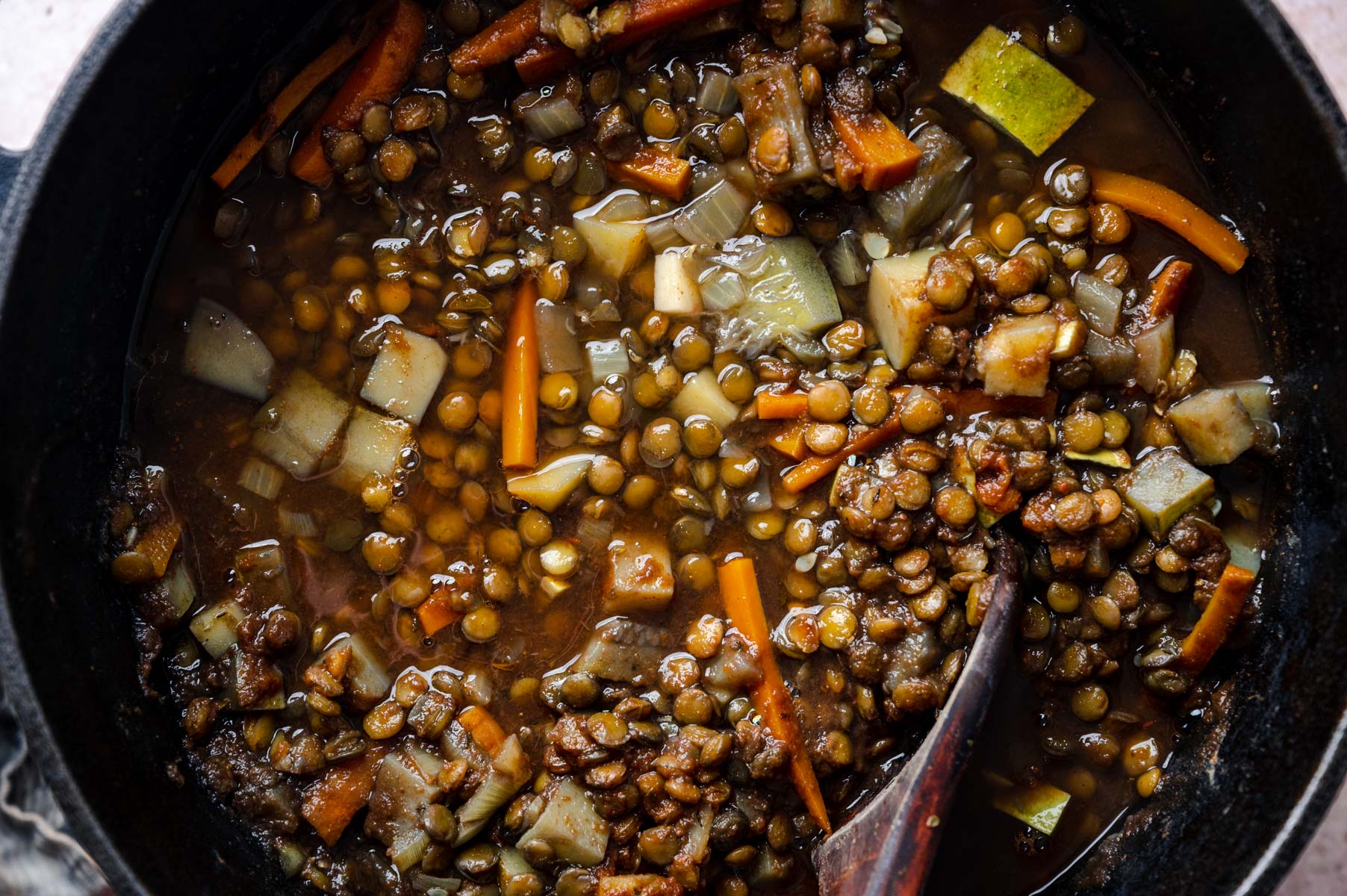 A close-up of a pot filled with a hearty lentil stew containing diced vegetables and carrots, stirred with a wooden spoon.