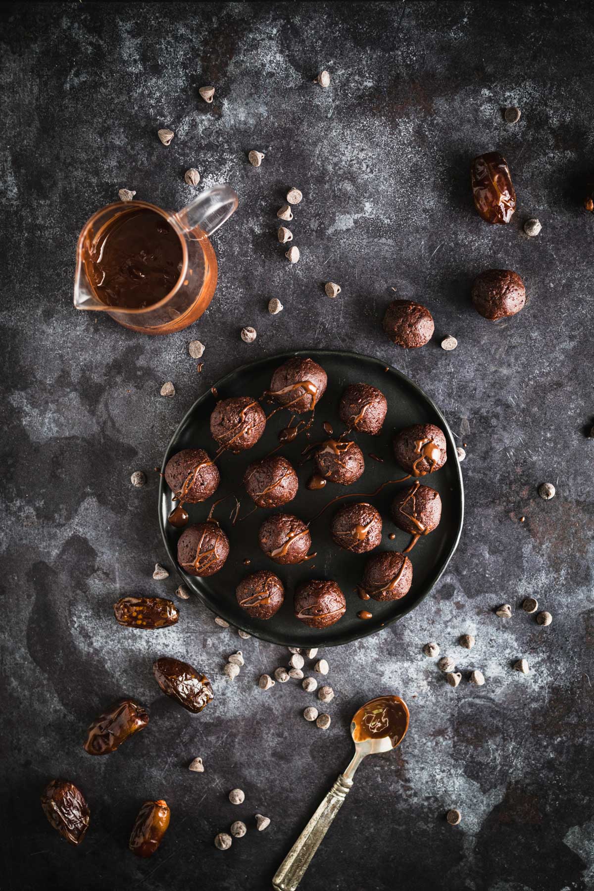 A plate of chocolate-covered date balls on a dark surface. A small pitcher filled with chocolate sauce, a spoon, and scattered dates and chocolate chips surround the plate.