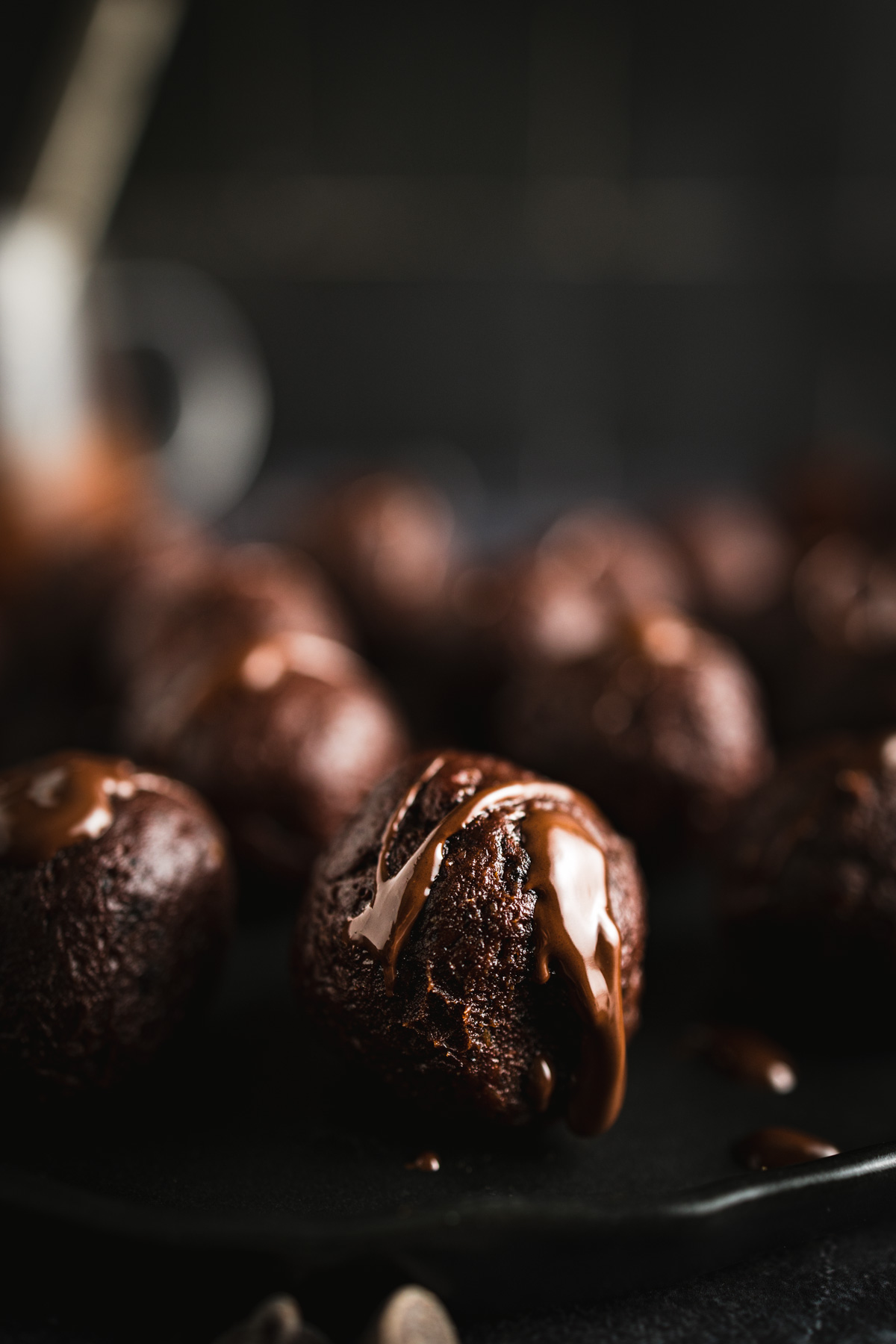 Close-up of chocolate truffles on a dark surface, each topped with a drizzle of melted chocolate. The background is blurred.