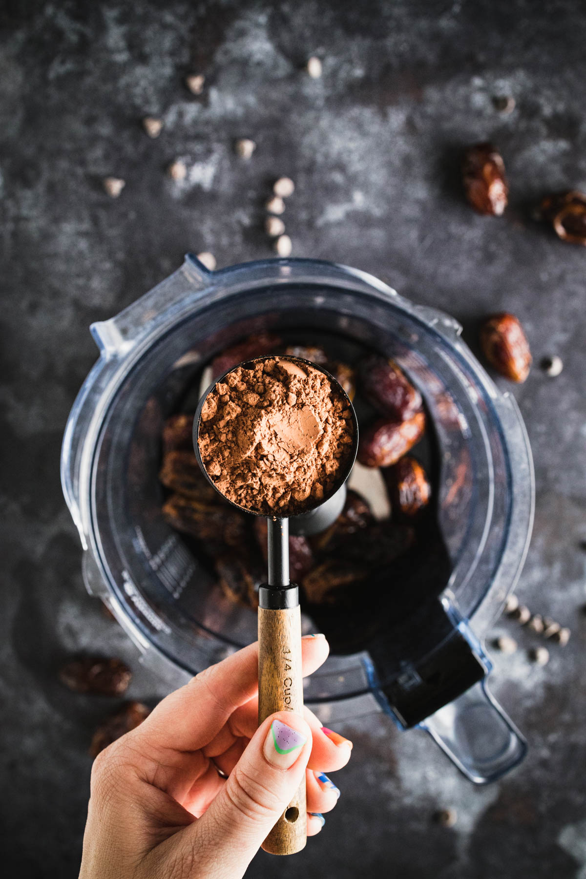 A hand holding a measuring spoon of cocoa powder above a blender filled with dates. Some dates and chocolate chips are scattered around the blender on a dark surface.