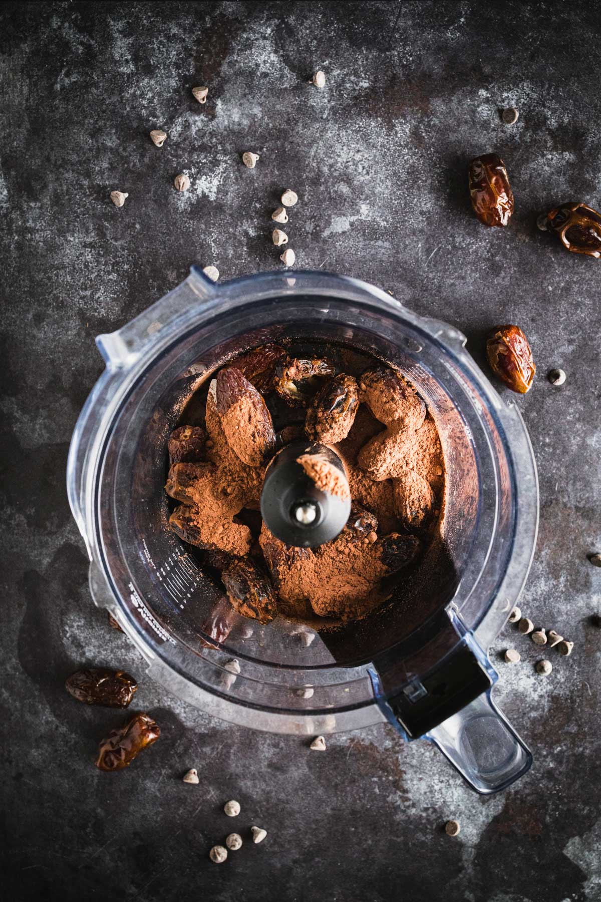 Top view of a food processor containing dates and cocoa powder, with some dates and chocolate chips scattered around on the dark surface.