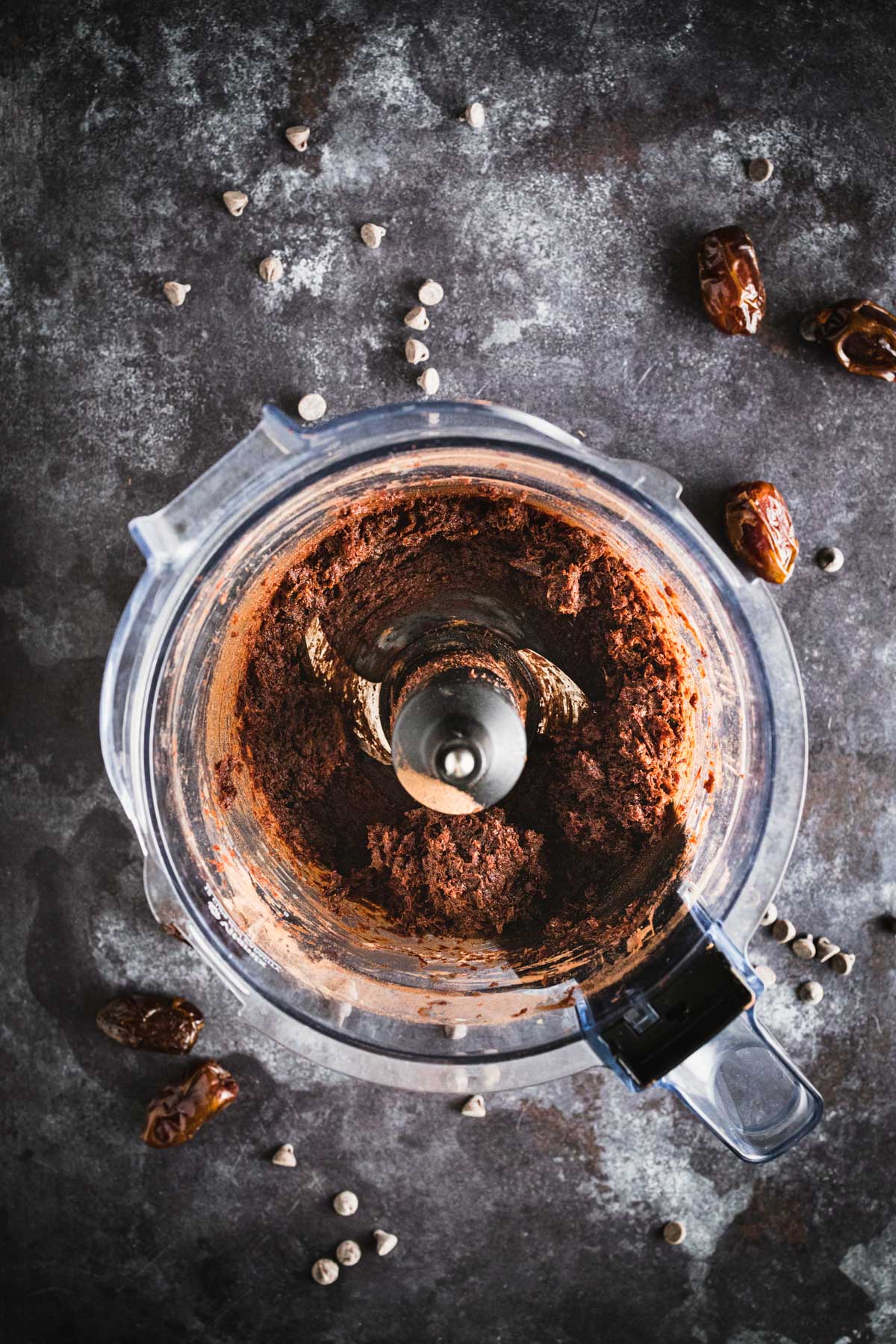 Top view of a food processor filled with blended chocolate mixture, surrounded by scattered date fruits and chocolate chips on a dark, textured surface.
