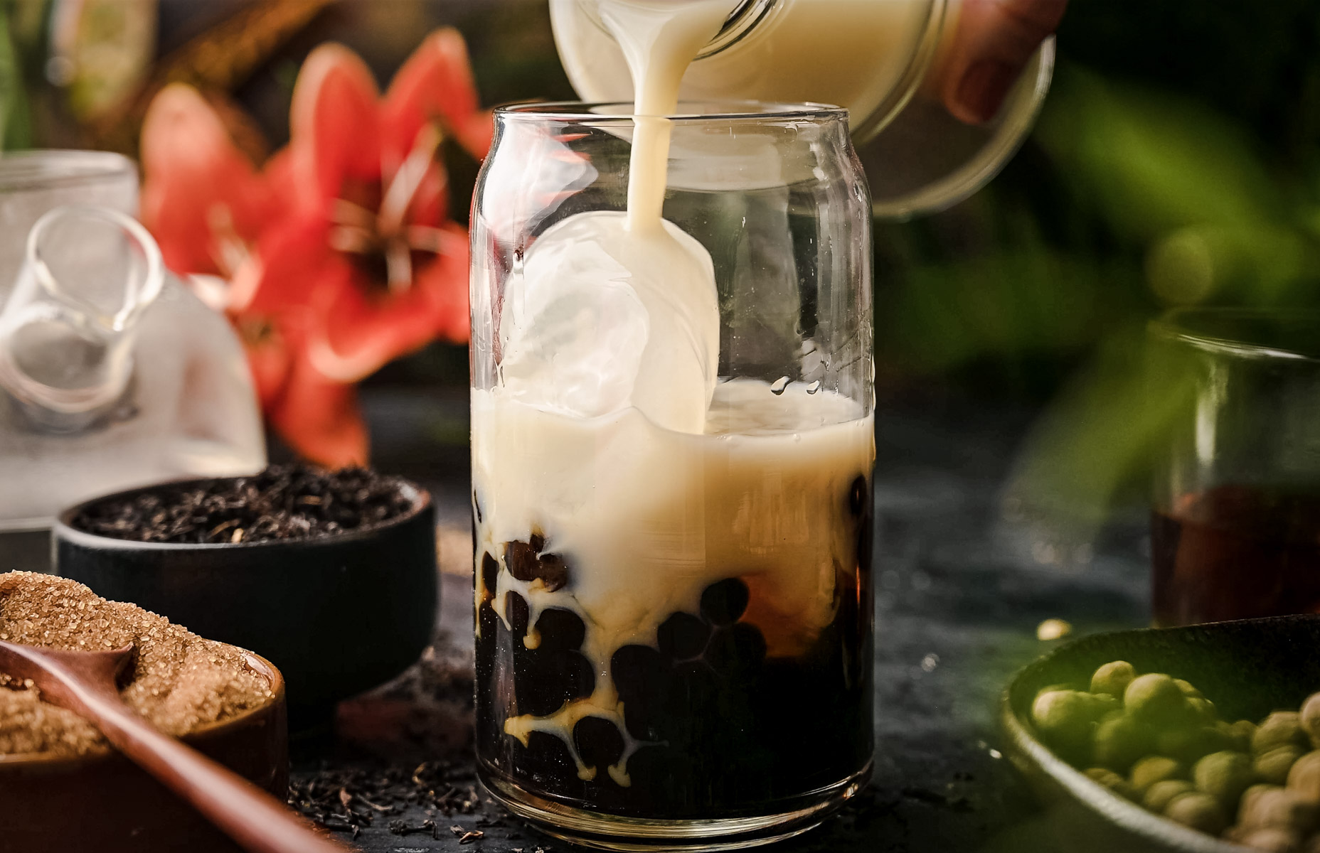 A glass jar being filled with milk over ice and black tapioca pearls, with sugar and tea leaves visible in the background.