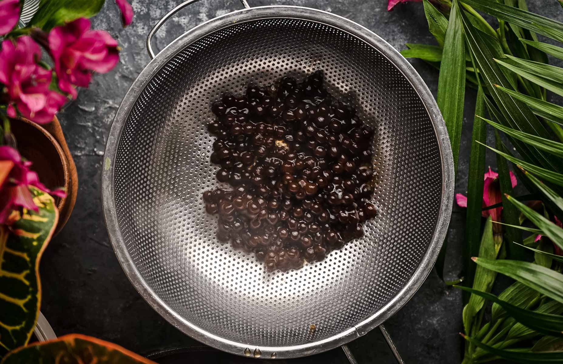 A metal strainer holding cooked tapioca pearls, surrounded by green leaves and pink flowers on a counter.