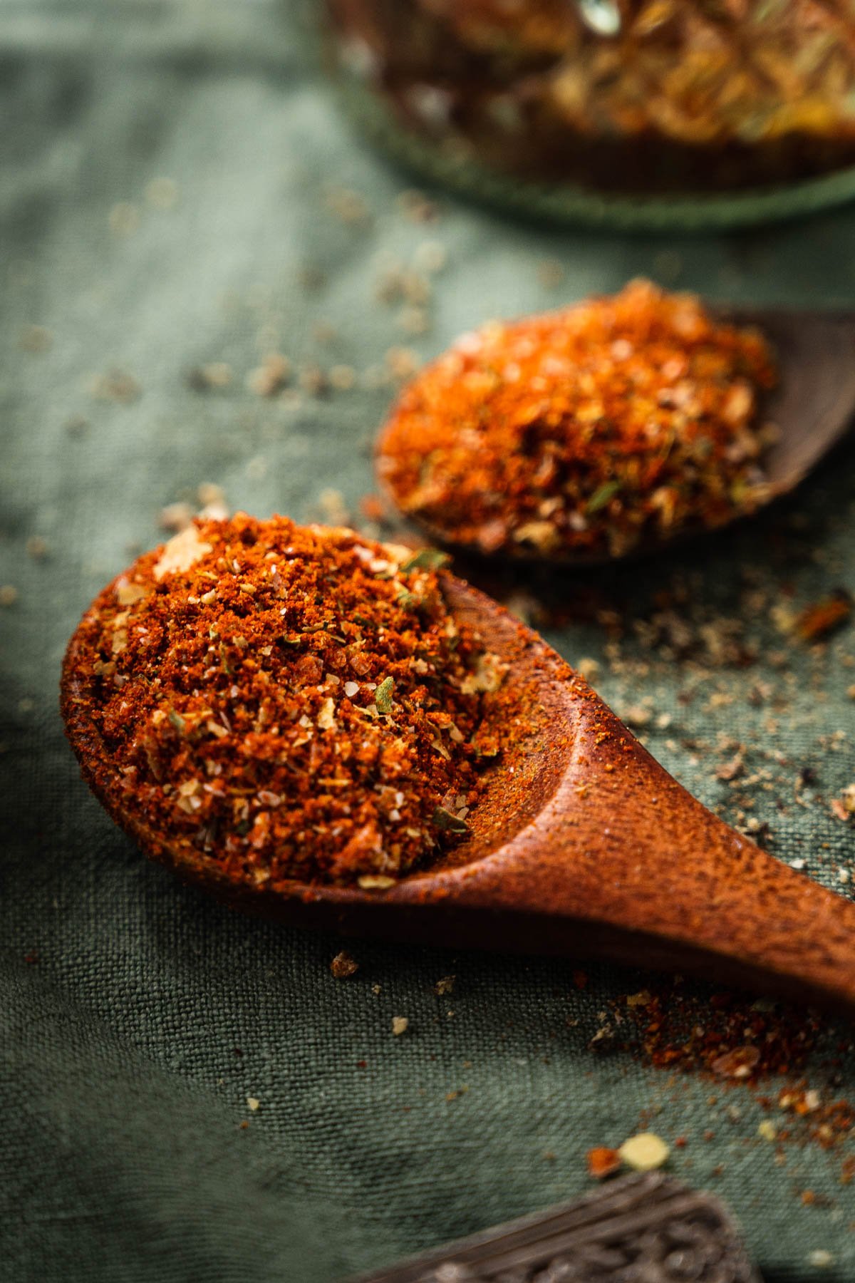 Close-up of two wooden spoons filled with a spice mix consisting of red and green dried herbs and seasonings, placed on a green fabric background.