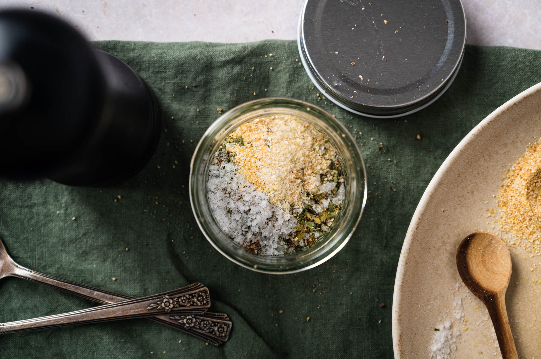 A jar of mixed seasonings, including salt and herbs, sits on a green cloth with a pepper grinder, silverware, and a partially visible plate and wooden spoon nearby.