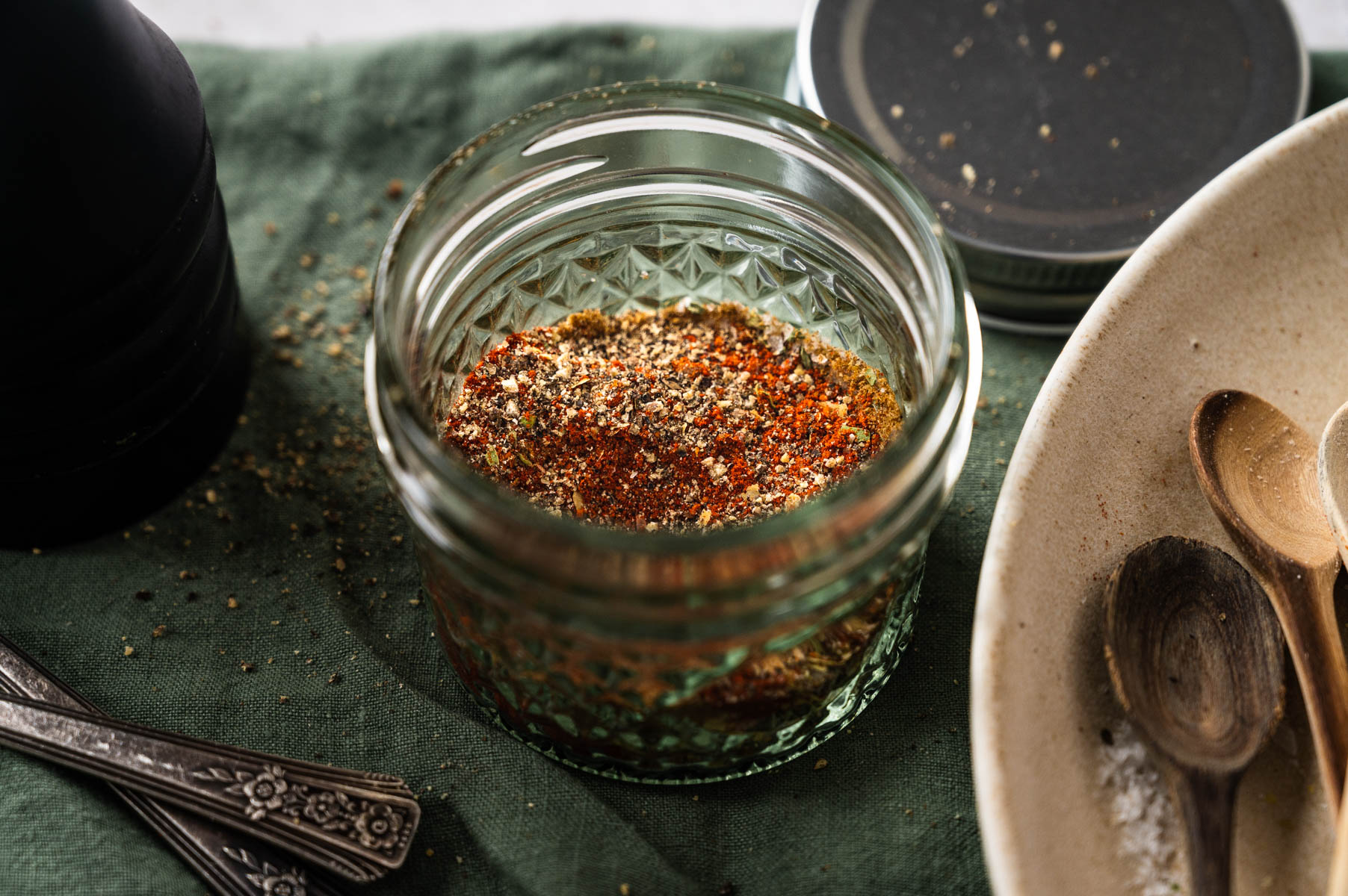 A small glass jar filled with a spice blend sits on a green cloth next to a lid, cutlery, and a bowl with wooden spoons.