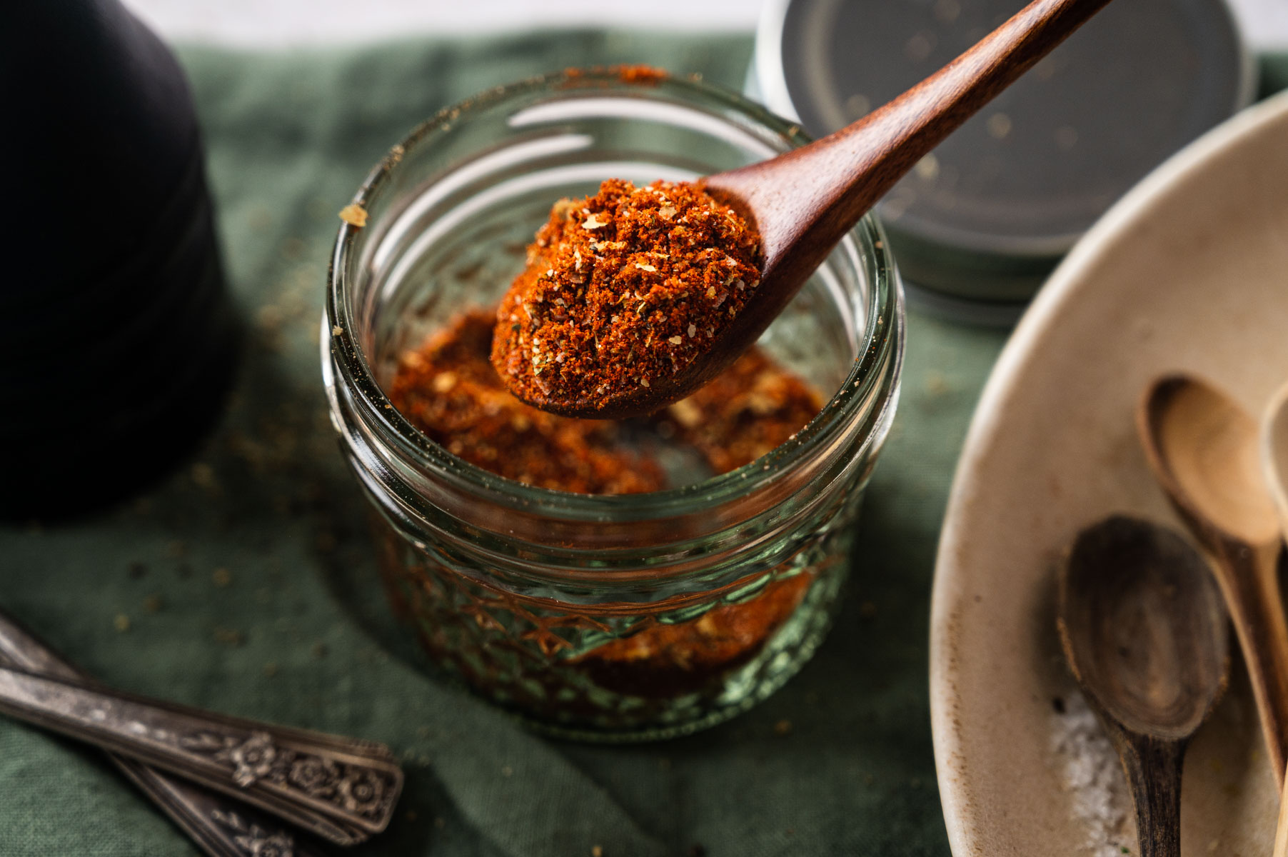 A wooden spoon holds red spice mixture above an open glass jar filled with the same spice. Spoons and a bowl are visible nearby on a green cloth.