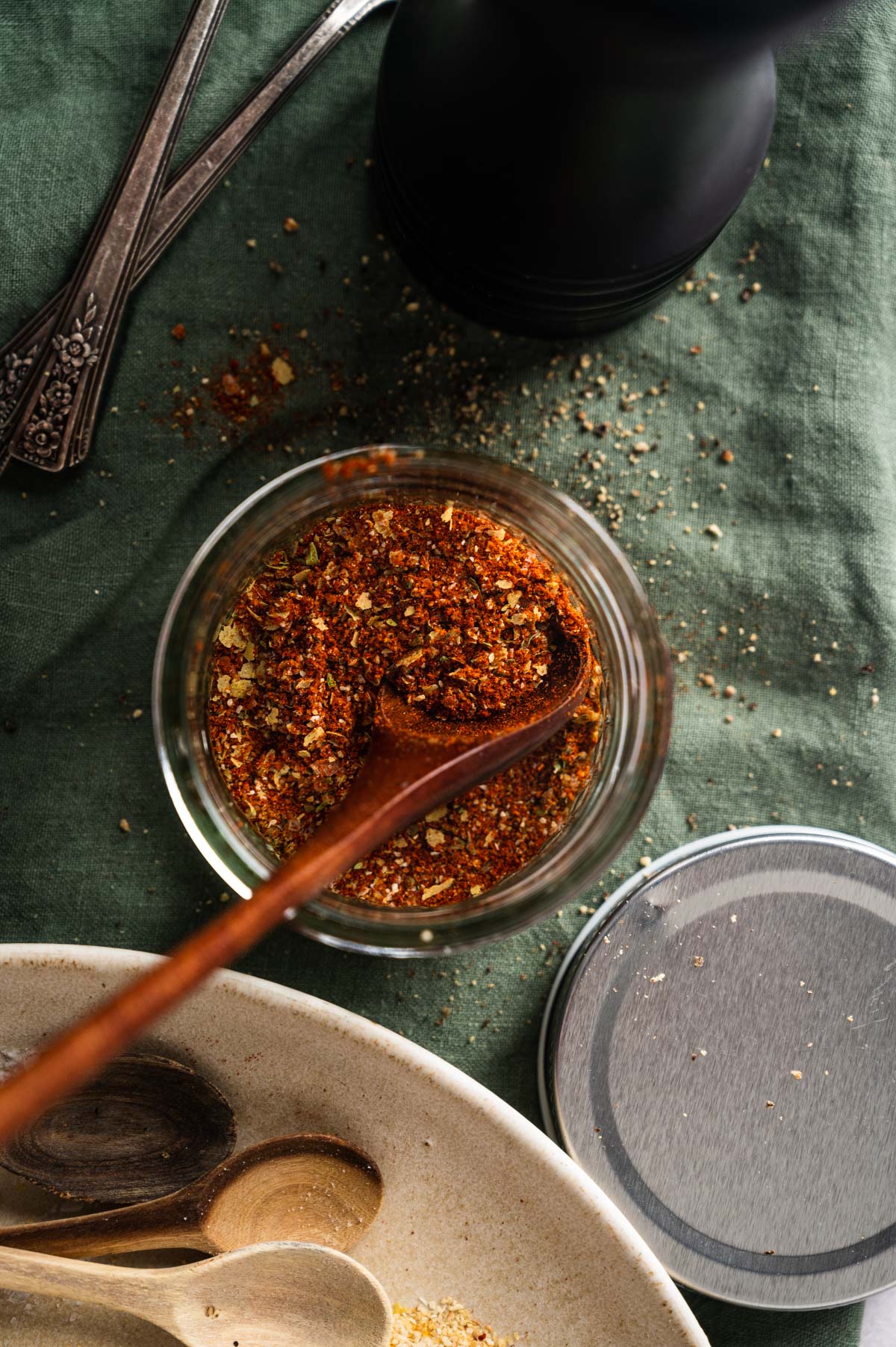 A top-down view of a jar filled with a homemade taco seasoning mix, a wooden spoon inside it, and surrounding utensils on a green cloth backdrop.