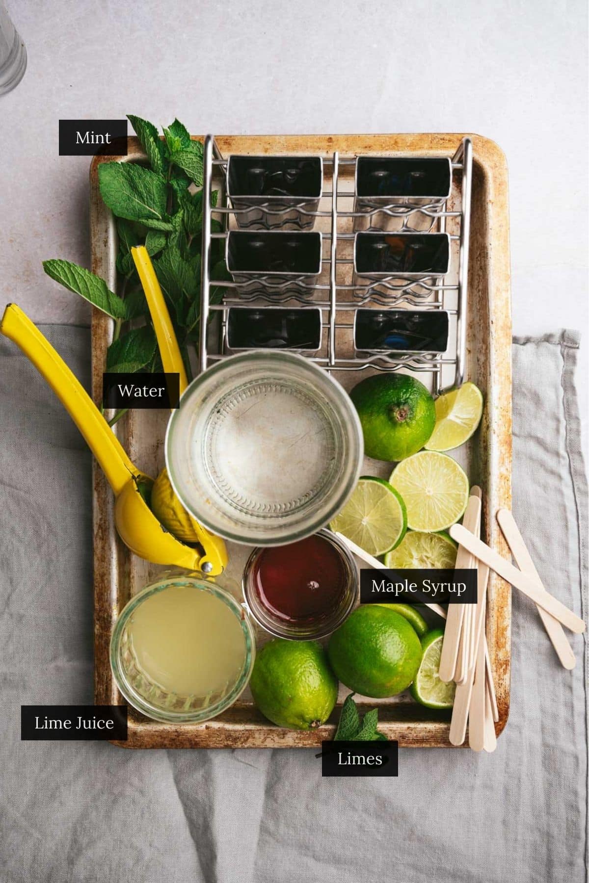 A tray holds mint leaves, a cup of water, maple syrup, lime juice for making lime popsicles, sliced limes, and metal popsicles molds.