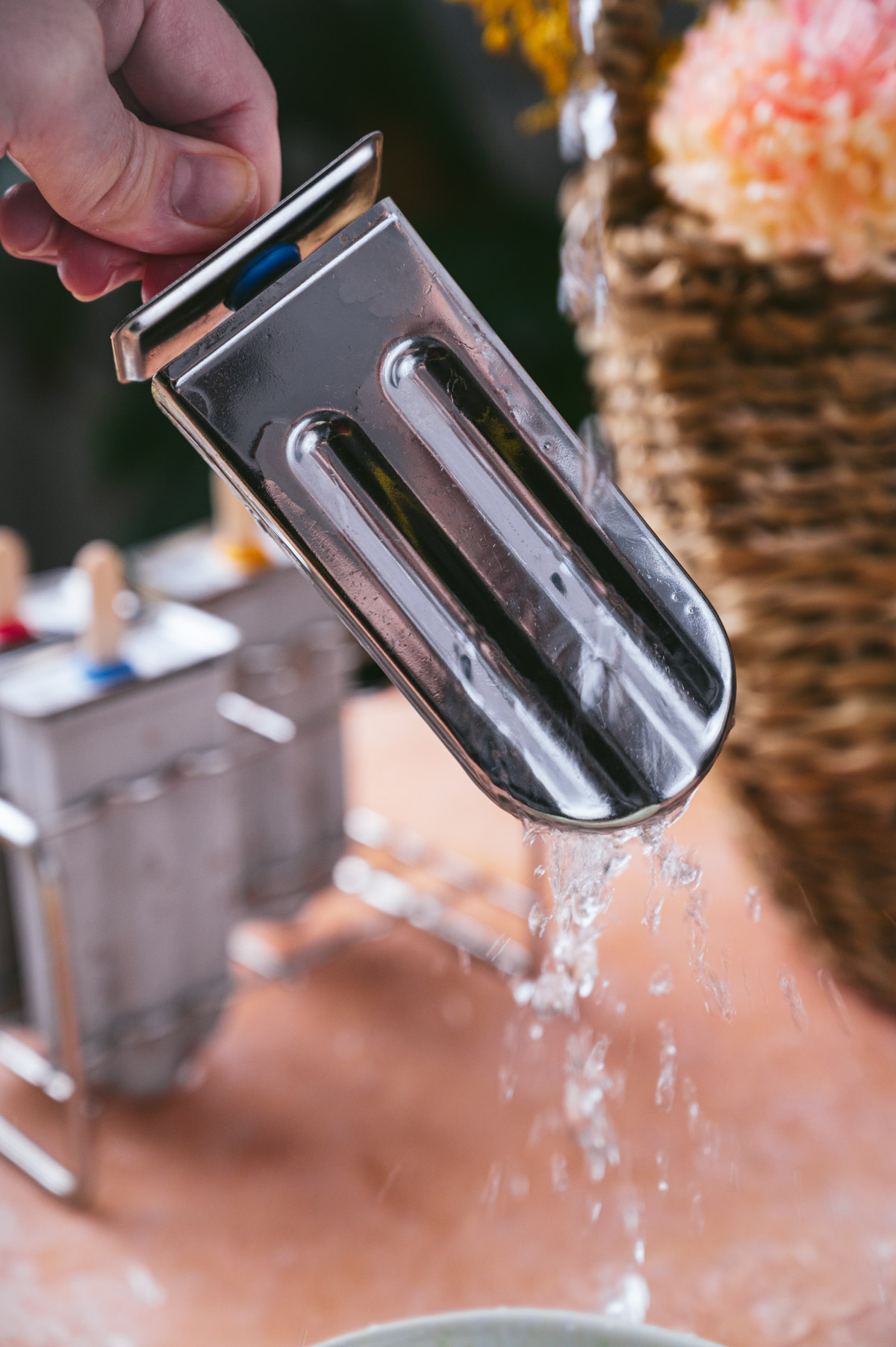 A hand holding a metallic popsicle mold with water dripping from it, with a wicker basket and popsicle molds in the background.