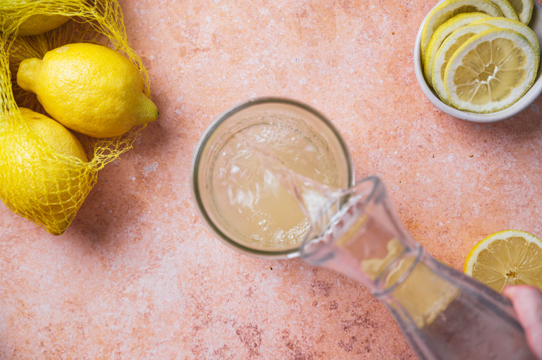 Top view of a glass being filled with lemonade from a carafe next to whole and sliced lemons on a peach-colored surface.